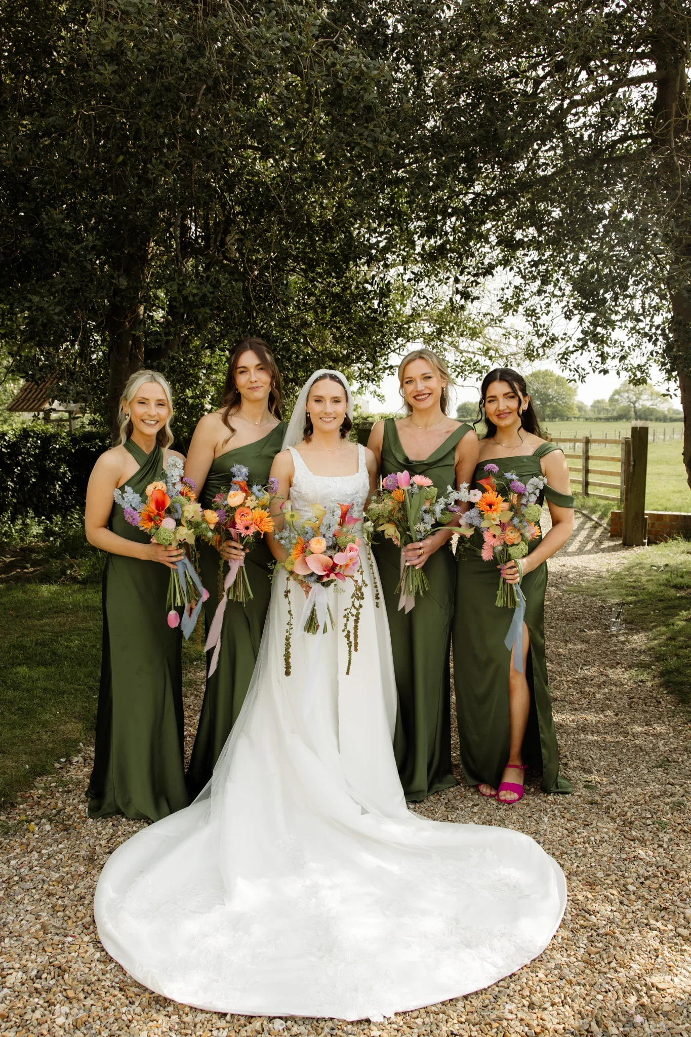 Five women dressed in green bridesmaid dresses and a bride in a white wedding gown holding colorful bouquets stand outdoors under a leafy tree. The scene appears to be a wedding celebration in a rural or countryside setting.