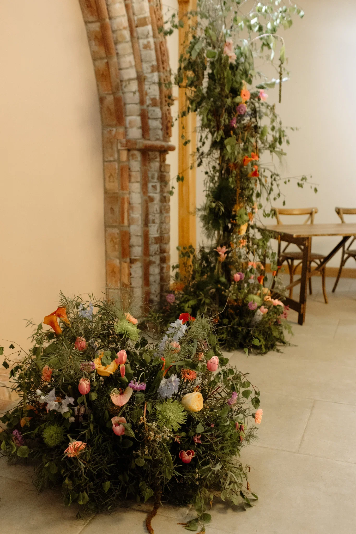 A large arrangement of various colorful flowers and greenery placed on the floor in front of a brick fireplace with a wooden mantel, next to a wooden dining table and chairs.