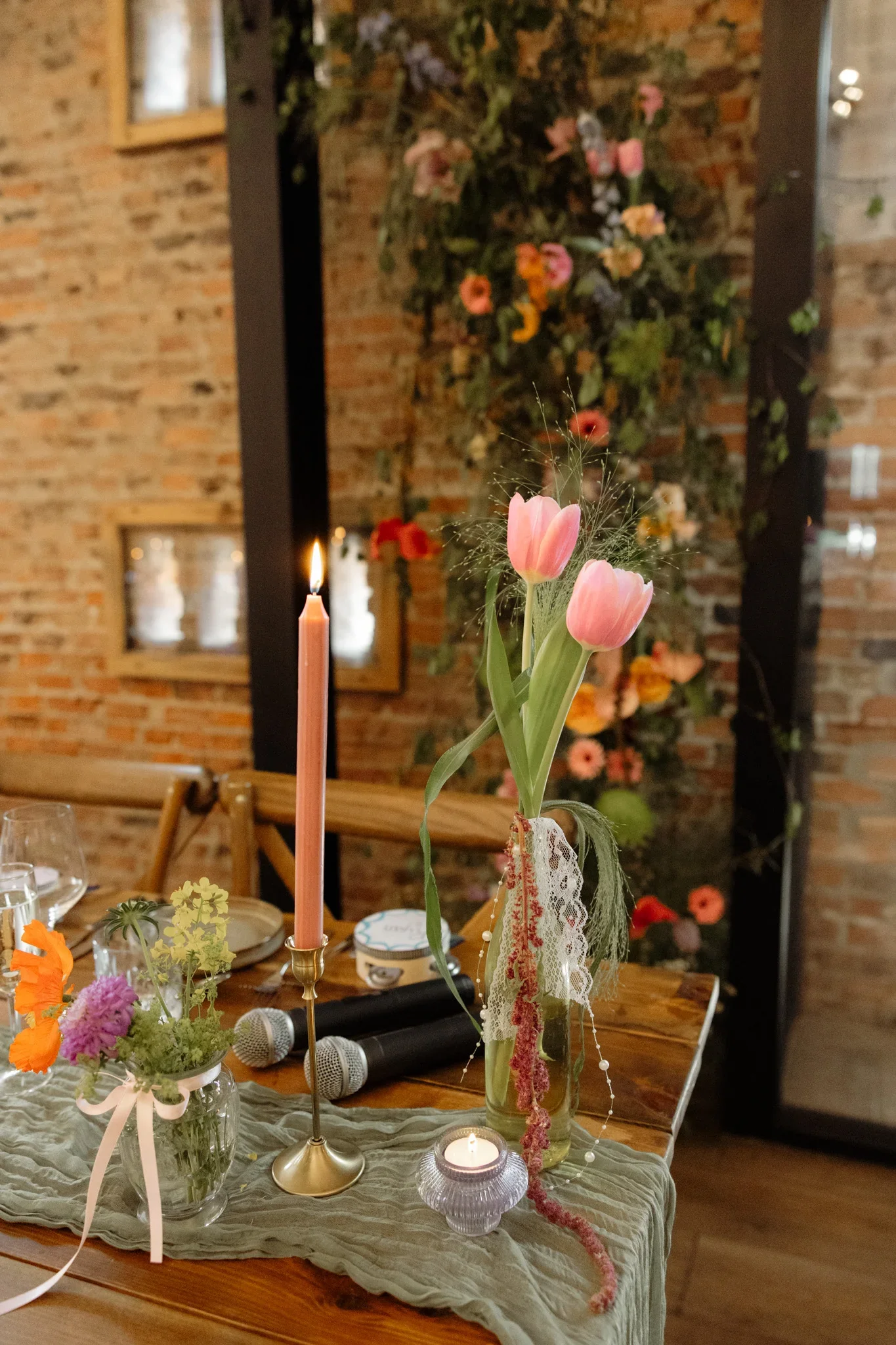 A table decorated with pink tulips in a glass vase, surrounded by candles, smaller flower arrangements, microphones, and various decorative items, set against a backdrop of a brick wall with floral decorations.
