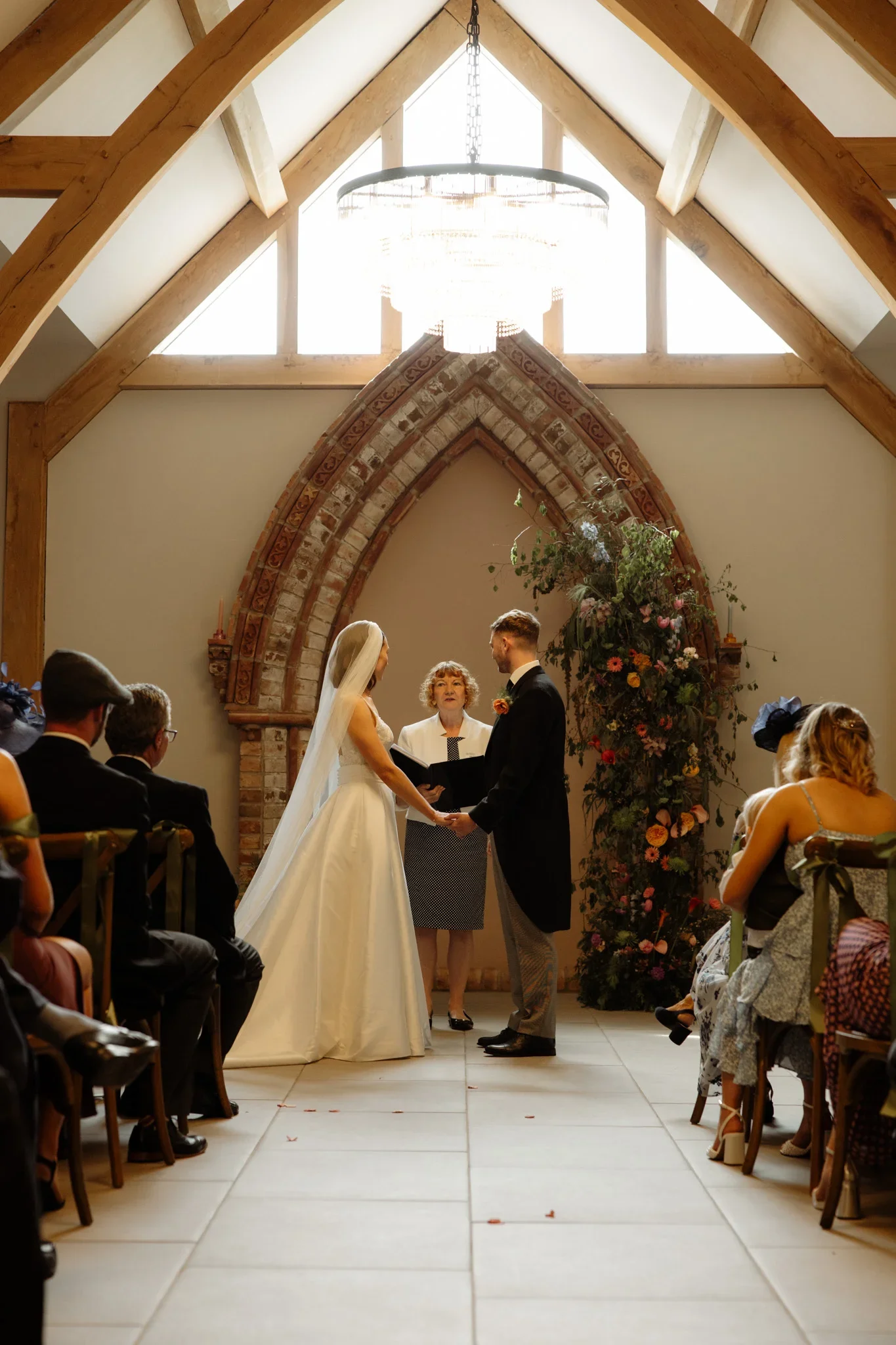 A wedding ceremony inside a church with wooden beams, a large chandelier, and a floral arch. The bride and groom are holding hands, facing each other, with a officiant between them. Guests are seated on either side, watching the ceremony.