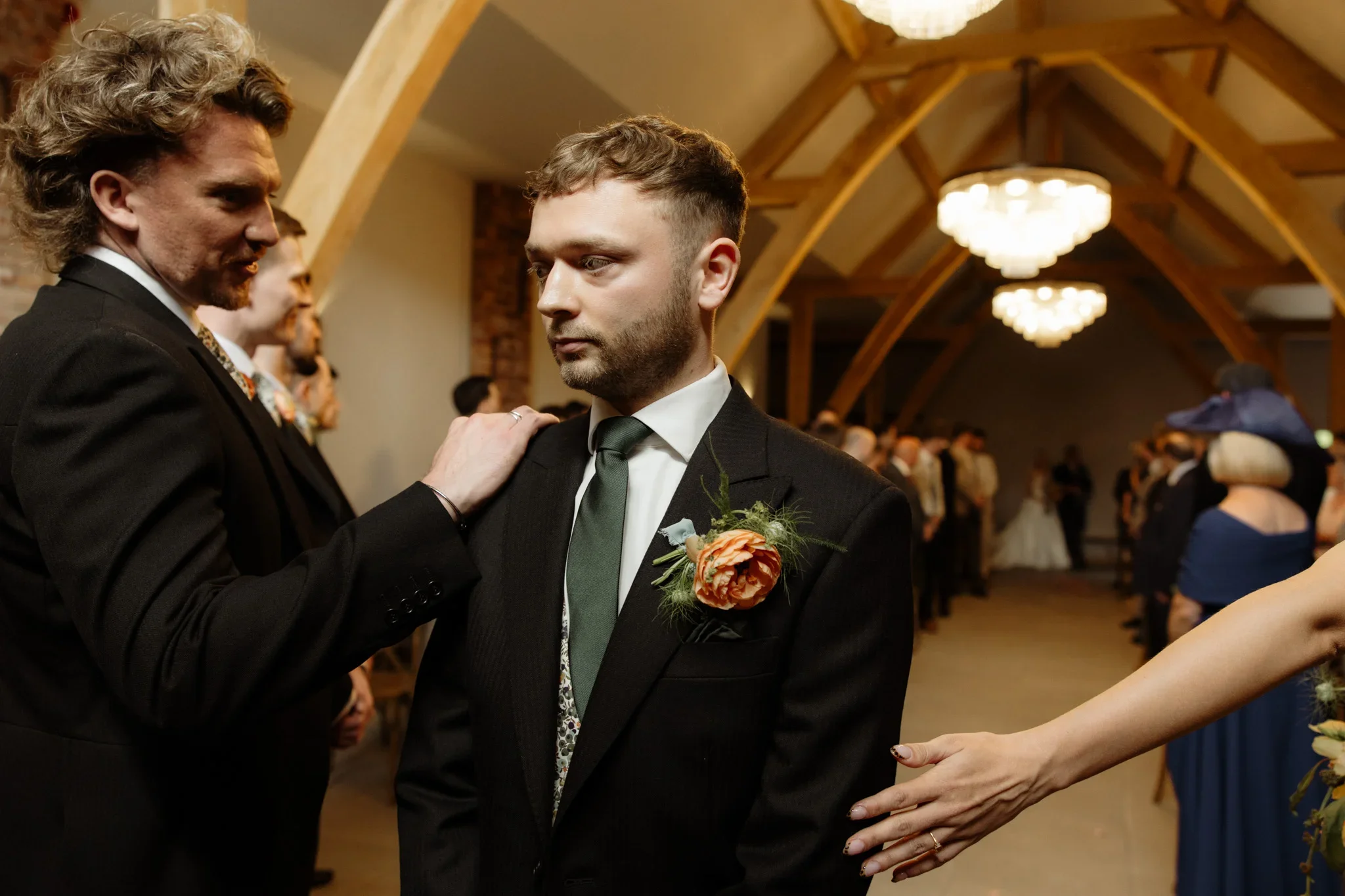 Two men in formal tuxedos, one with a boutonniere, holding hands and looking at each other during a wedding ceremony in a decorated hall with chandeliers, surrounded by guests.