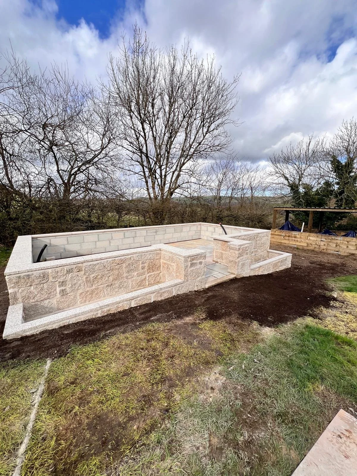 A stone fire pit under construction outdoors, with a backdrop of leafless trees and a partly cloudy sky.