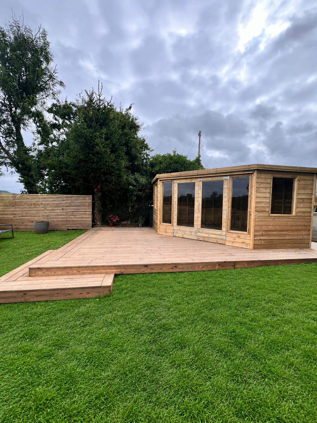 A newly built wooden deck and a small wooden garden house with glass doors in a backyard, with trees and cloudy sky in the background.