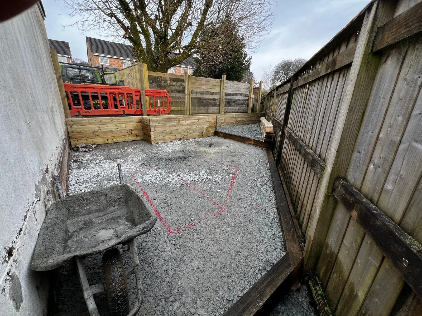 A small backyard under construction with a gravel ground, a wheelbarrow, wooden fencing, and red and black outdoor equipment stacked against the fence.