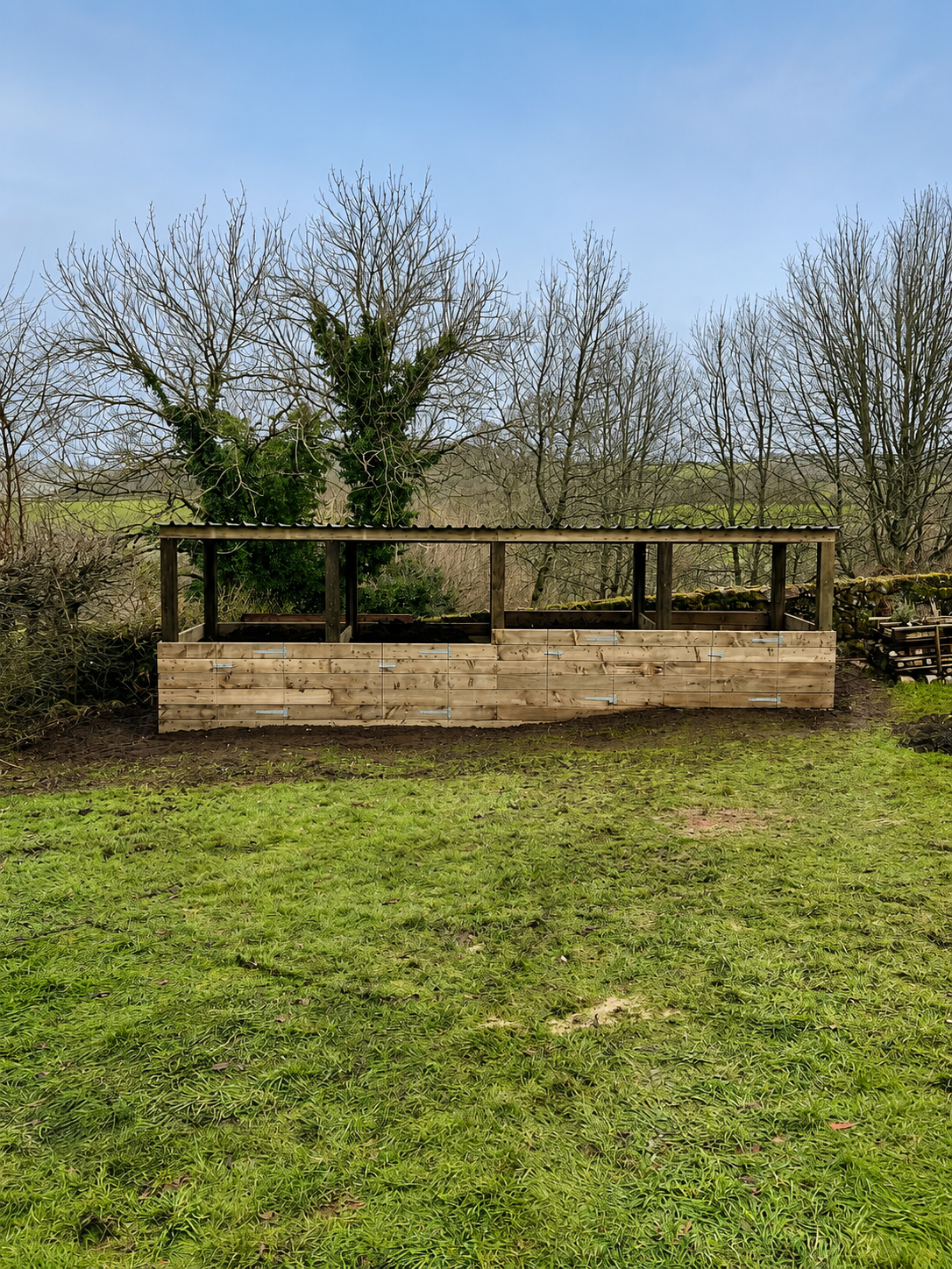 A wooden outdoor structure under construction in a backyard with green grass, leafless trees, and a blue sky.