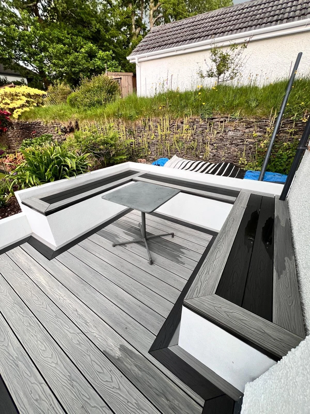 A modern outdoor patio with light gray wooden decking, built-in white and black seating, a small table, and garden plants and bushes in the background.