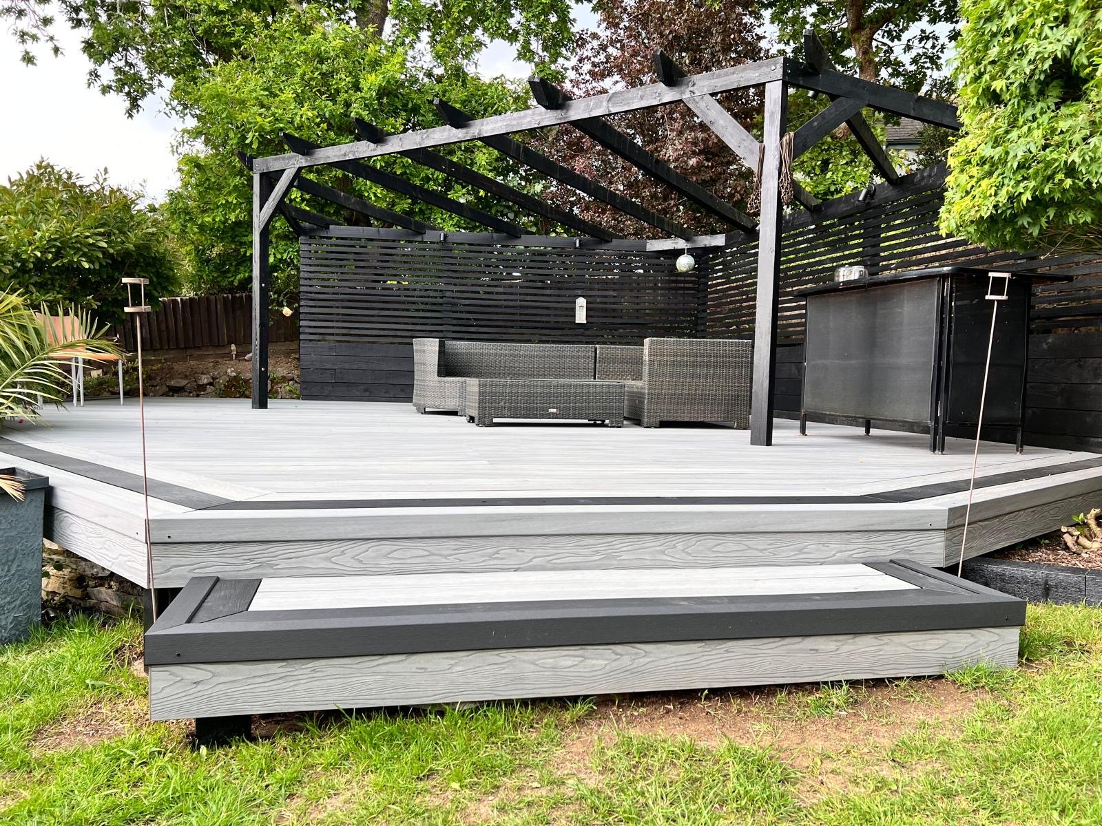 A modern outdoor deck with black and gray furniture, including a sectional sofa, a bar cart, and a pergola structure surrounded by green trees.