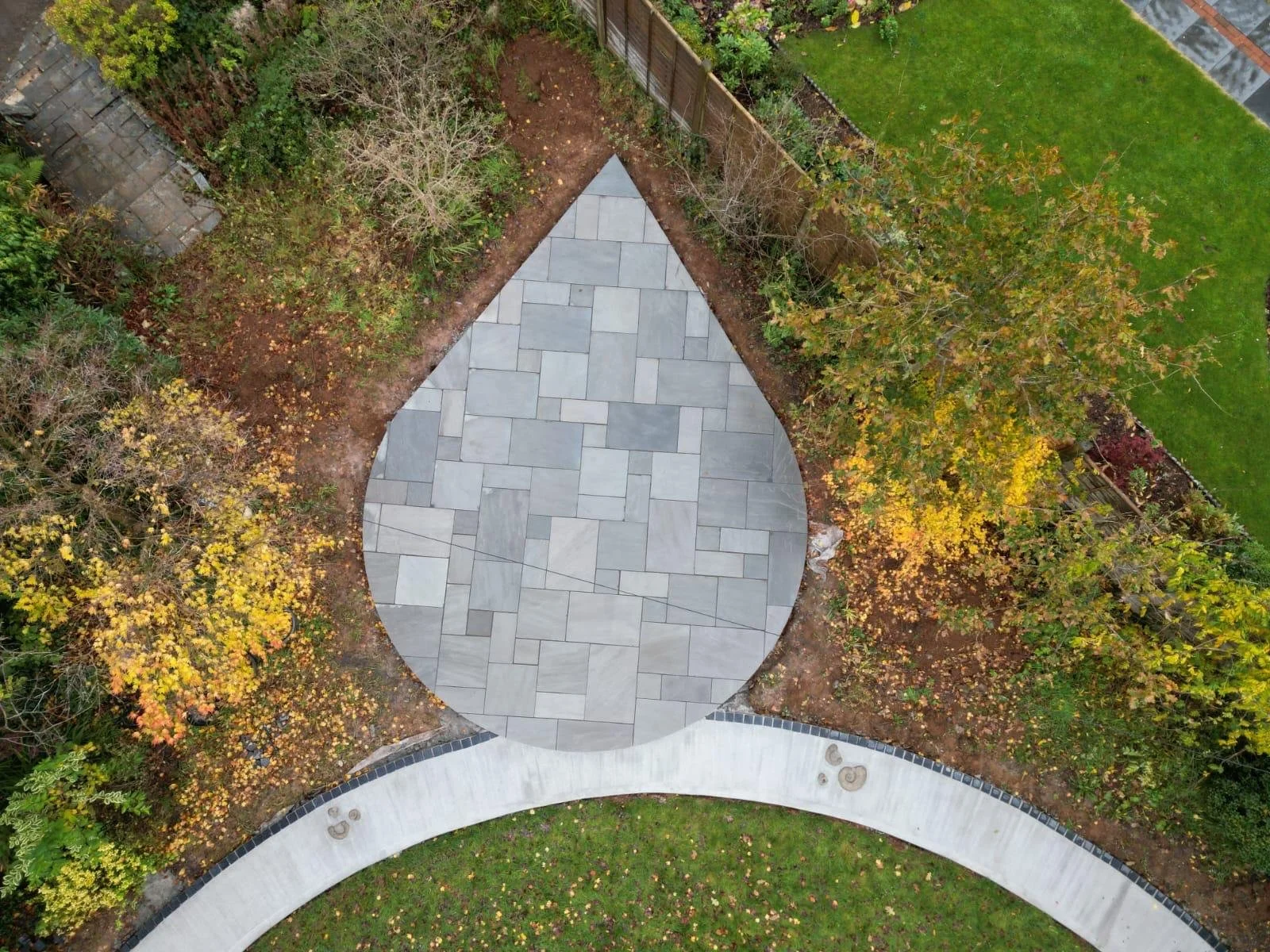 An aerial view of a backyard with a rounded, paved patio surrounded by leafless and leafy trees, a green lawn, and garden beds along a wooden fence.