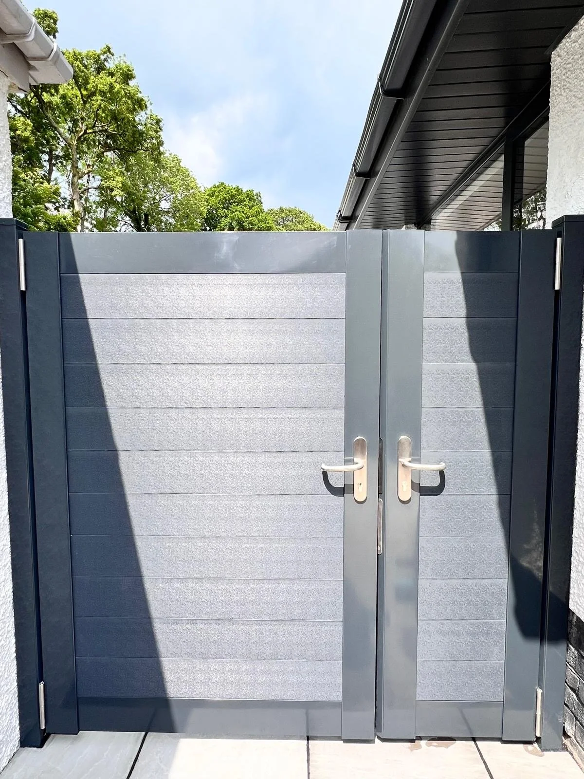 A gray metal double gate with handles, part of a modern fence, attached to black posts. The background includes trees, a blue sky, and part of a house with black roofing and white exterior walls.