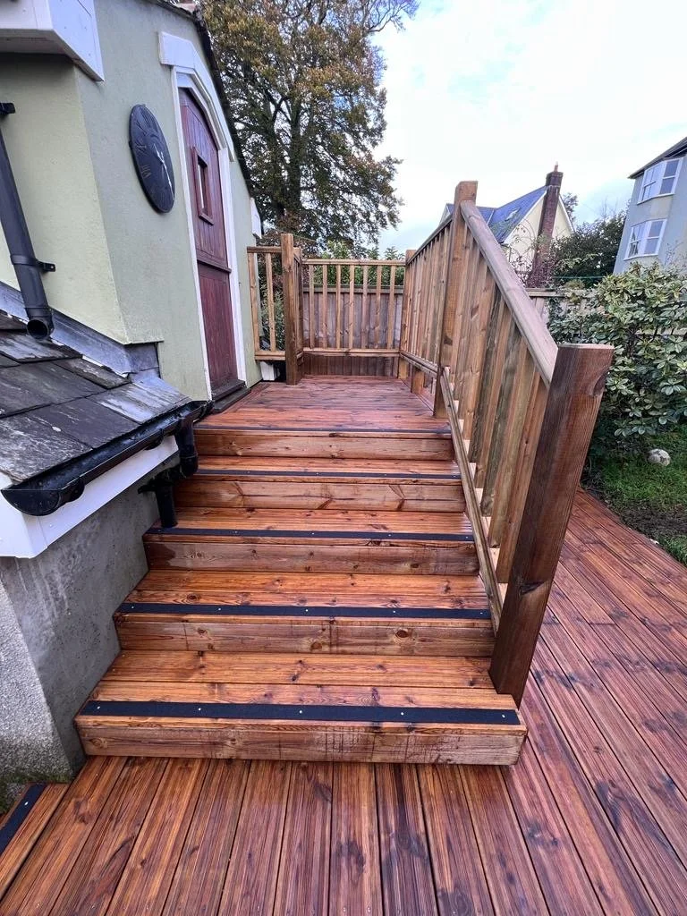 Newly built wooden deck with stairs and railing attached to a house, with neighboring houses and trees in the background.