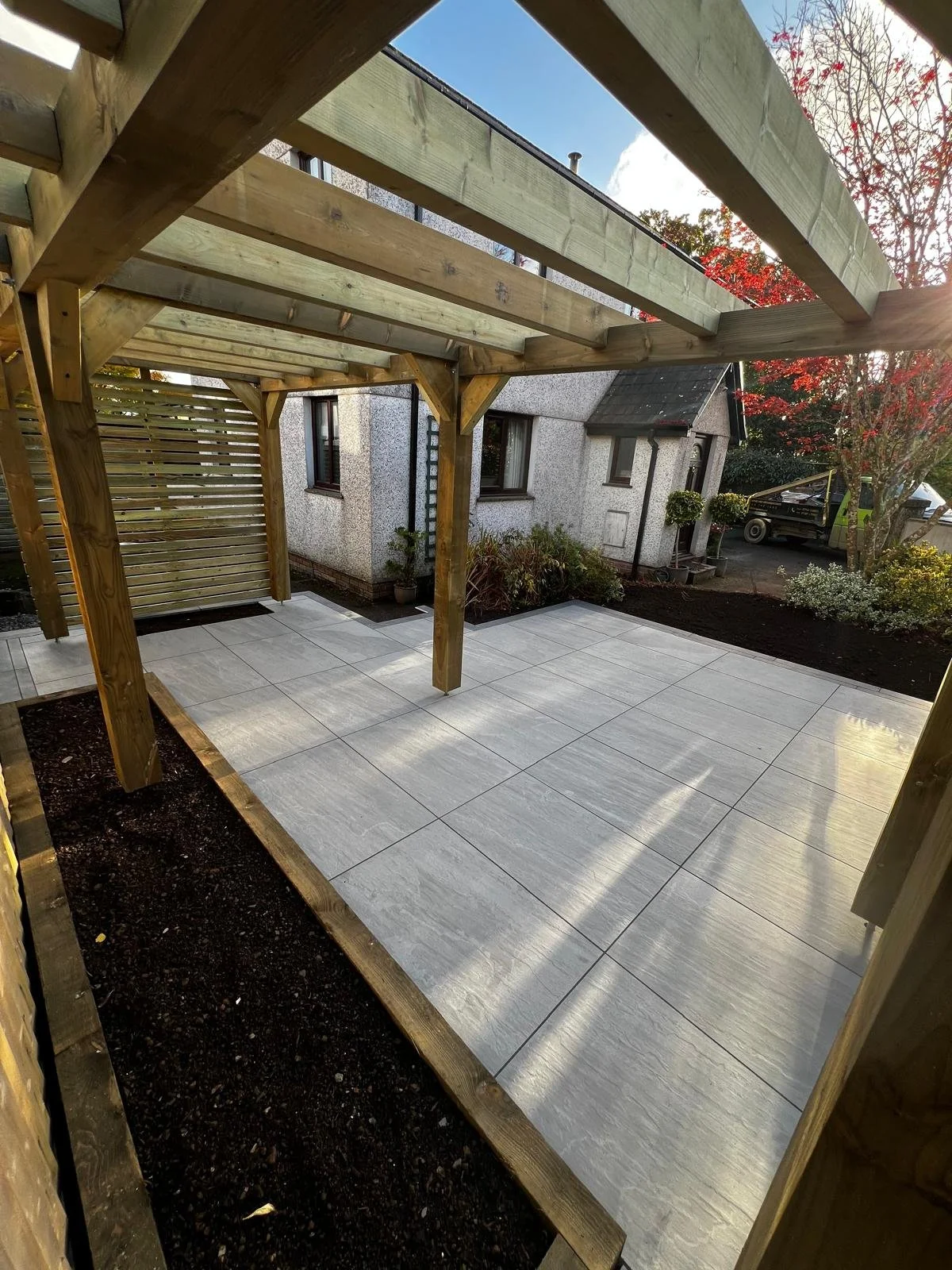 A backyard patio area with large grey tiles, a wooden pergola framework, and surrounding plants and garden.