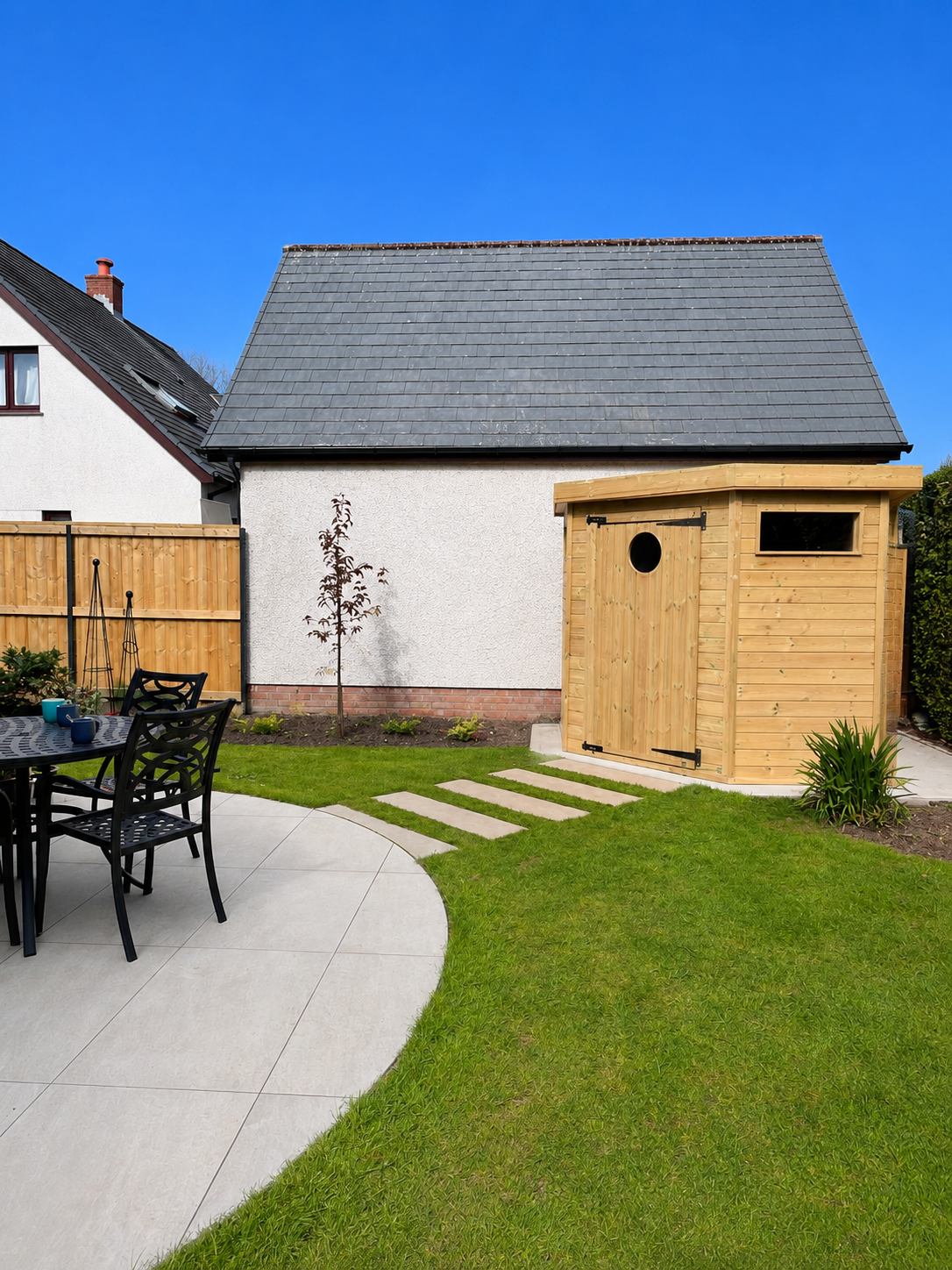 Backyard with a wooden shed, a small tree, a patio with chairs and table, and a green lawn under a clear blue sky.