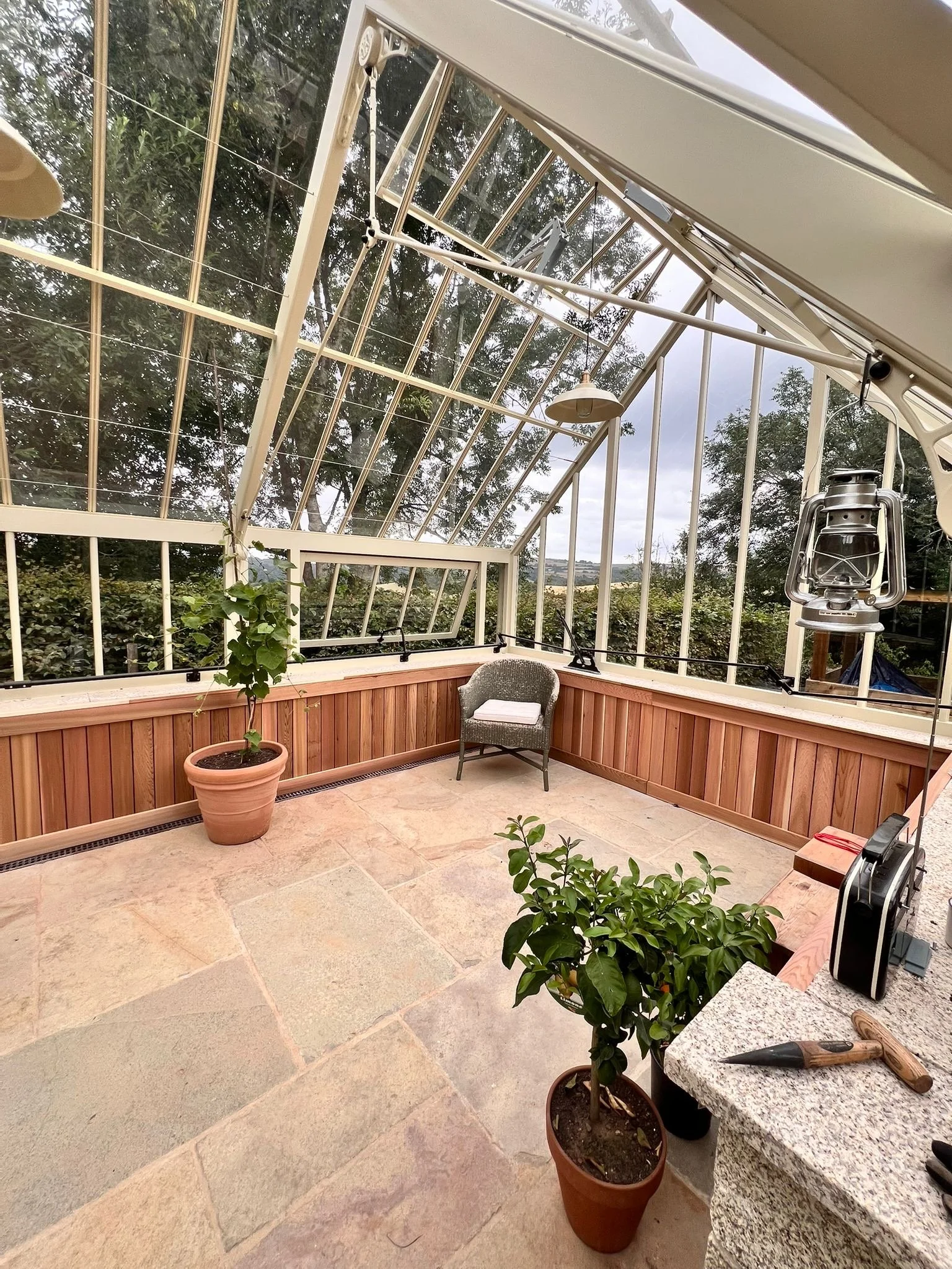 A greenhouse with a glass roof and walls, containing potted plants, a gray chair with a white cushion, hanging lights, outside view of trees, and a granite countertop with a pen on it.