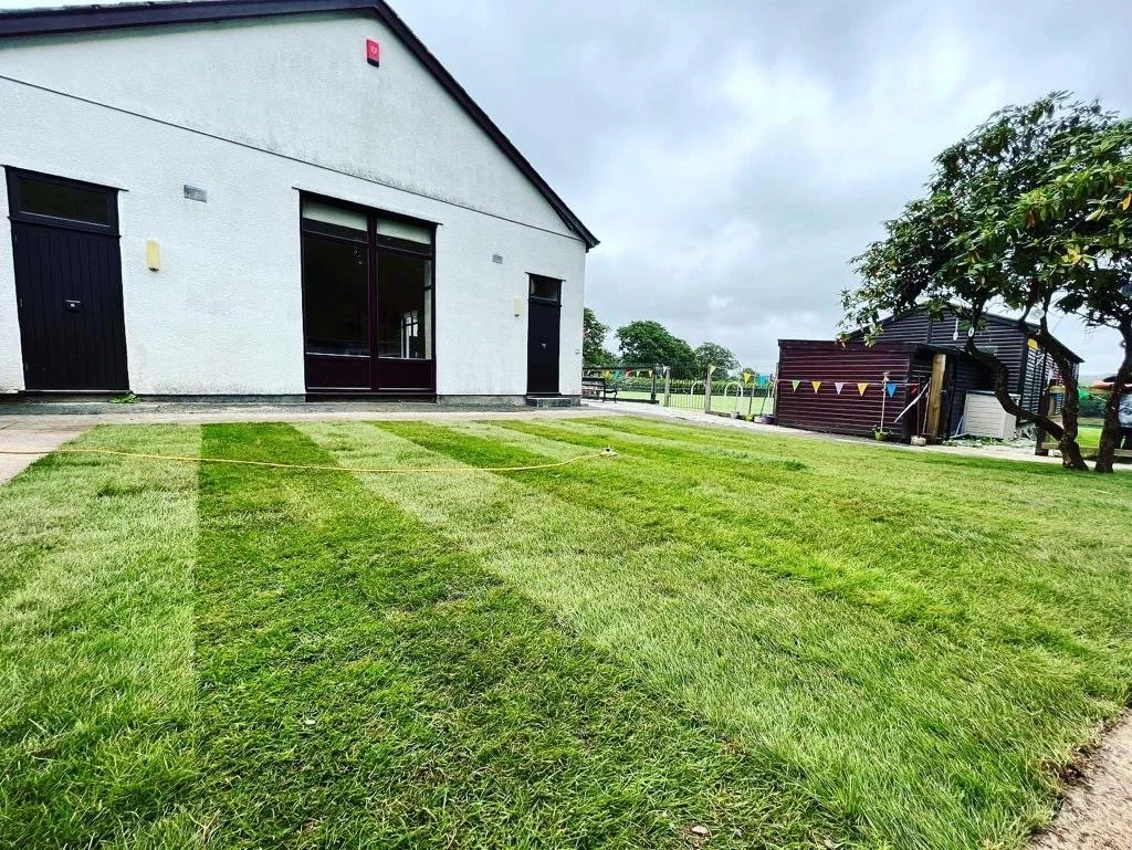 A building with a white exterior wall, a large window, and black doors, next to a well-maintained grassy area with patterned mowing lines, and a small wooden shed decorated with bunting and surrounded by trees.