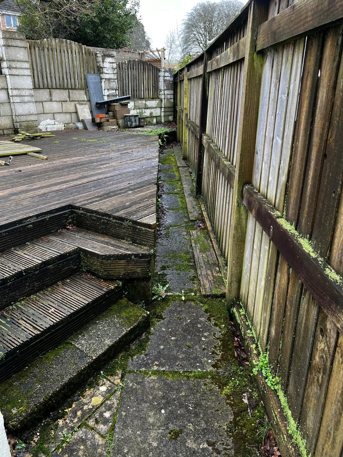 Backyard with moss-covered stone pathway and wooden deck with steps, surrounded by weathered wooden and concrete block fences, some gardening tools and plants in the corner.