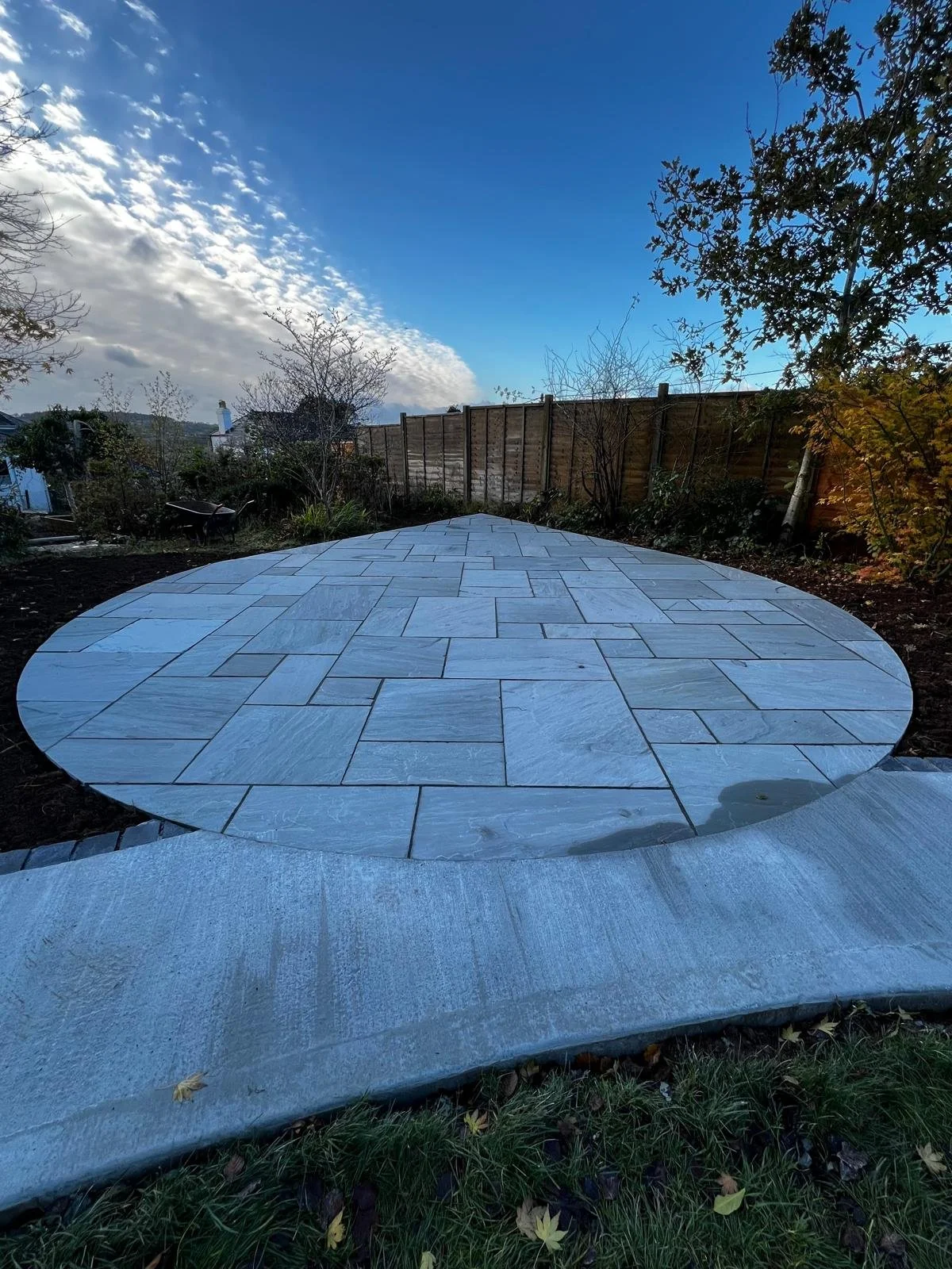 A newly constructed round stone patio with a curving edge in a backyard, bordered by a wooden fence and trees under a partly cloudy sky.