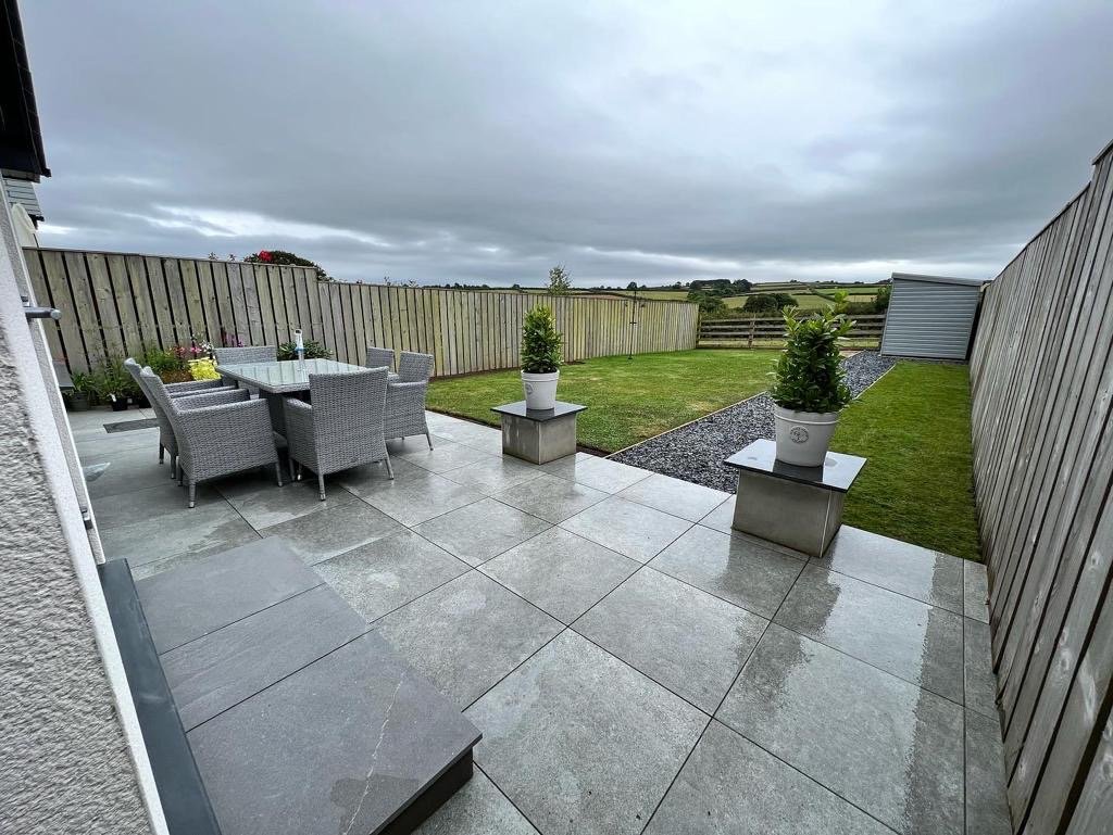 Cloudy backyard with tiled patio, outdoor dining table with six chairs, two potted plants on concrete pedestals, small lawn, gravel pathway, and wooden fence, with open field and cloudy sky in the background.