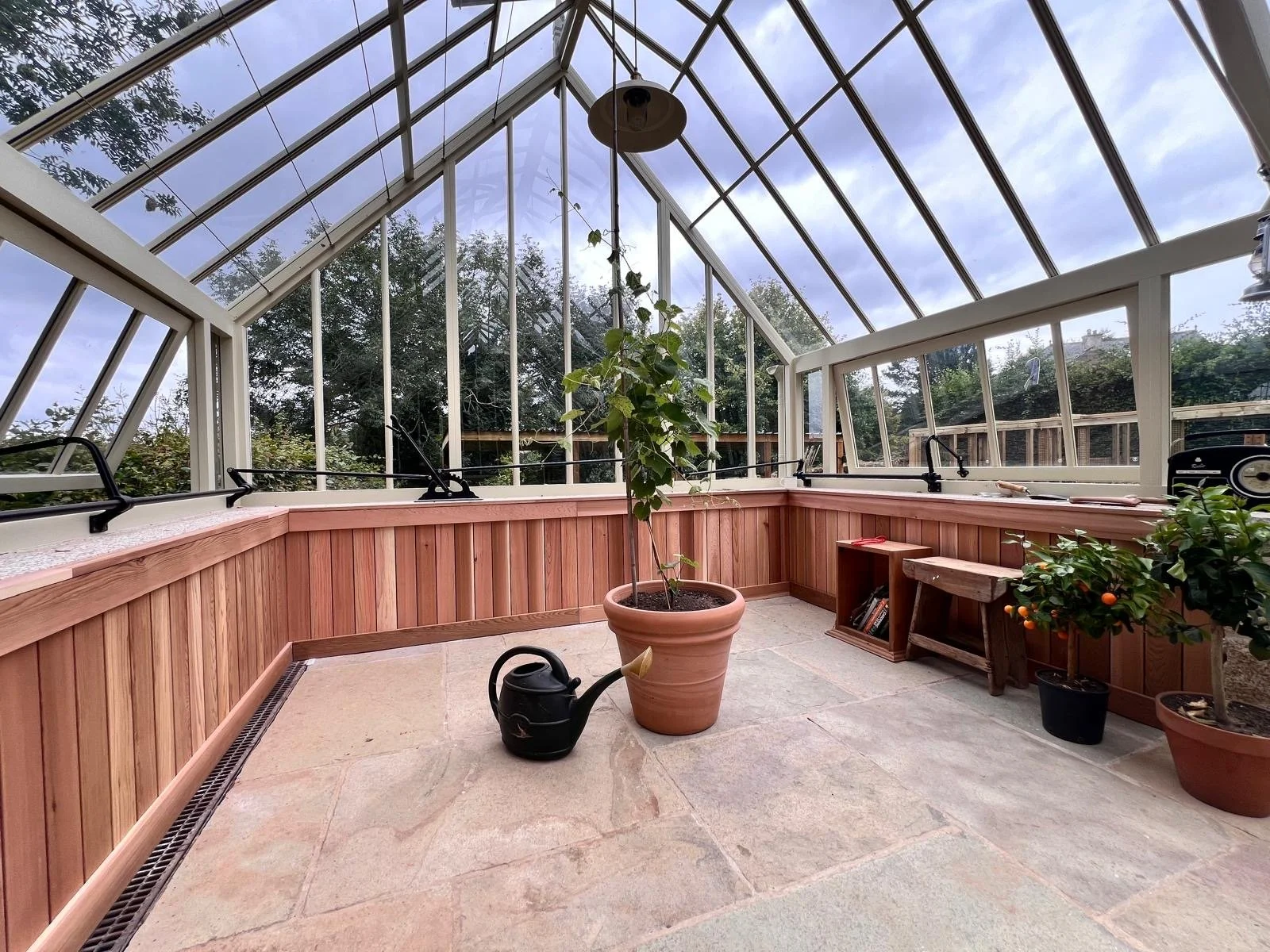 Inside a greenhouse with glass walls and ceiling, containing potted plants, a watering can, and wooden benches, with trees visible outside.
