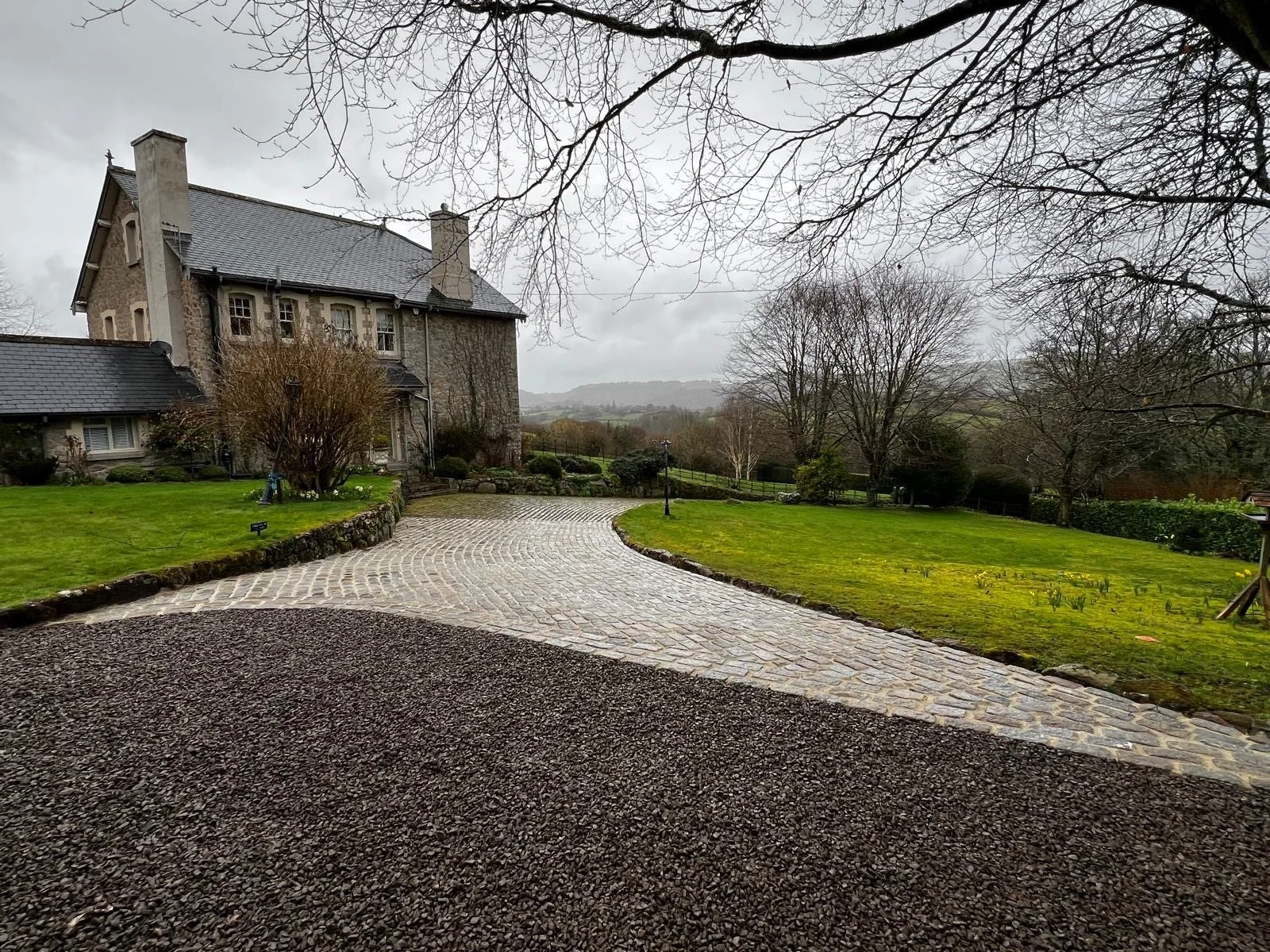 A stone driveway curves past a large lawn with leafless trees and a stone house in the background on a cloudy day.