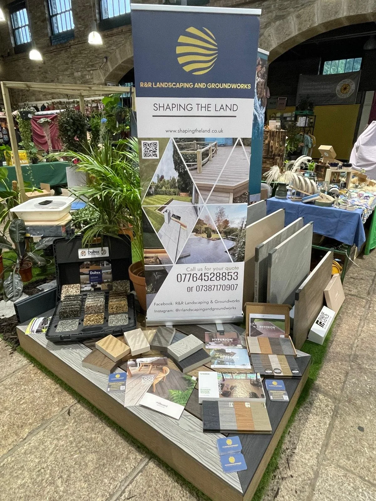 Display booth for R&R Landscaping and Groundworks at an indoor event, showcasing landscape paving materials, plants, and promotional materials. Banner with company logo, contact info, and images of projects.