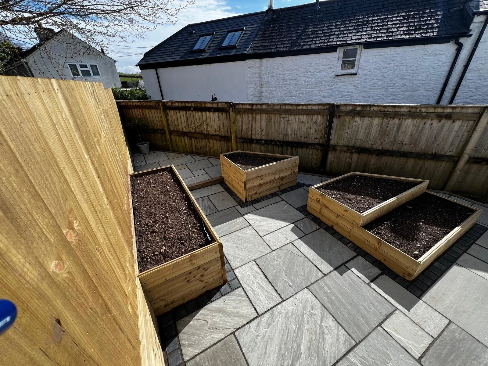 A small outdoor patio with four square wooden garden beds filled with soil, enclosed by a wooden fence, with a white house in the background under cloudy sky.
