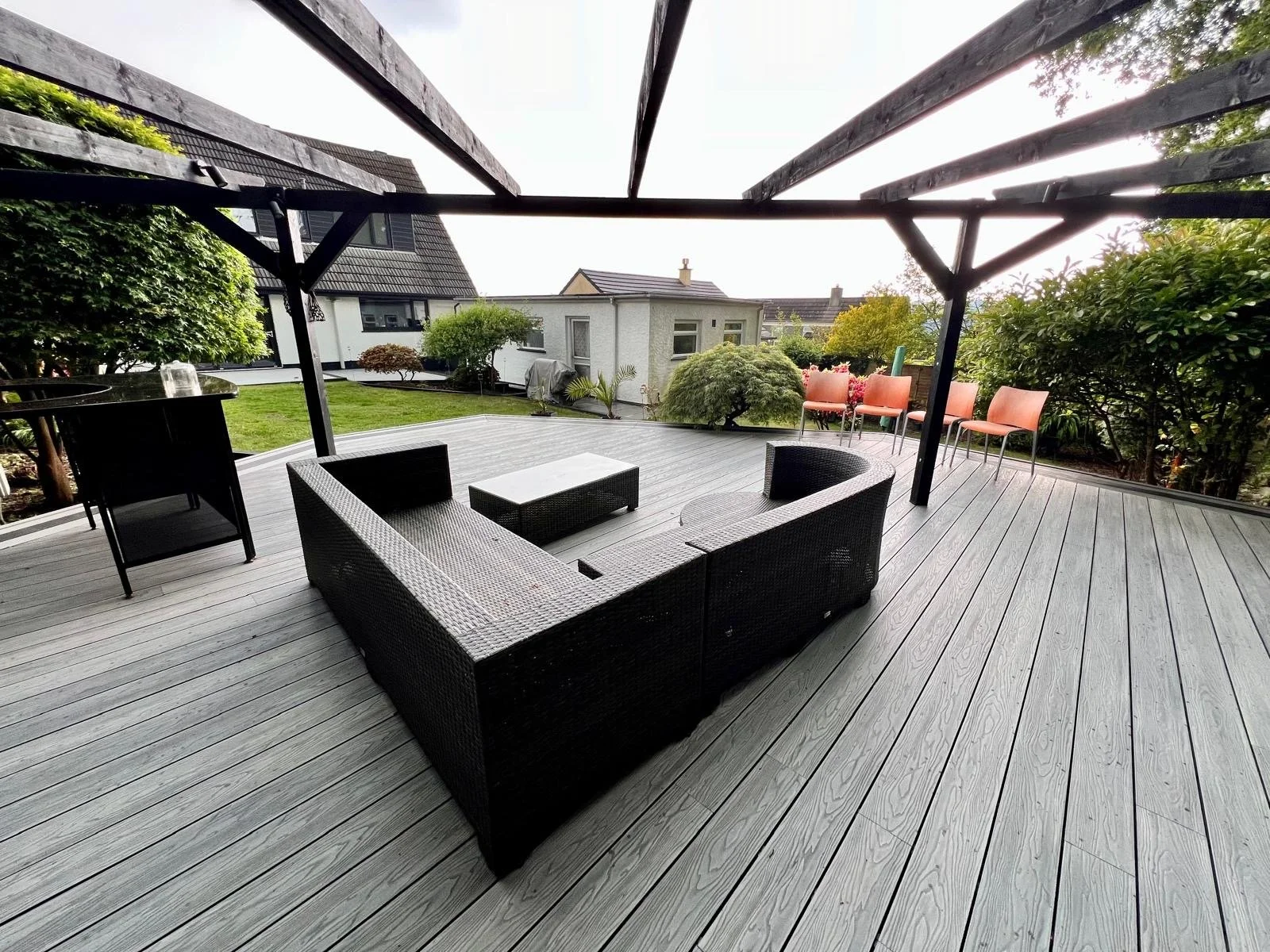 Spacious outdoor deck with black wicker sectional sofa, small white coffee table, and orange chairs in the background, surrounded by greenery and neighboring houses.