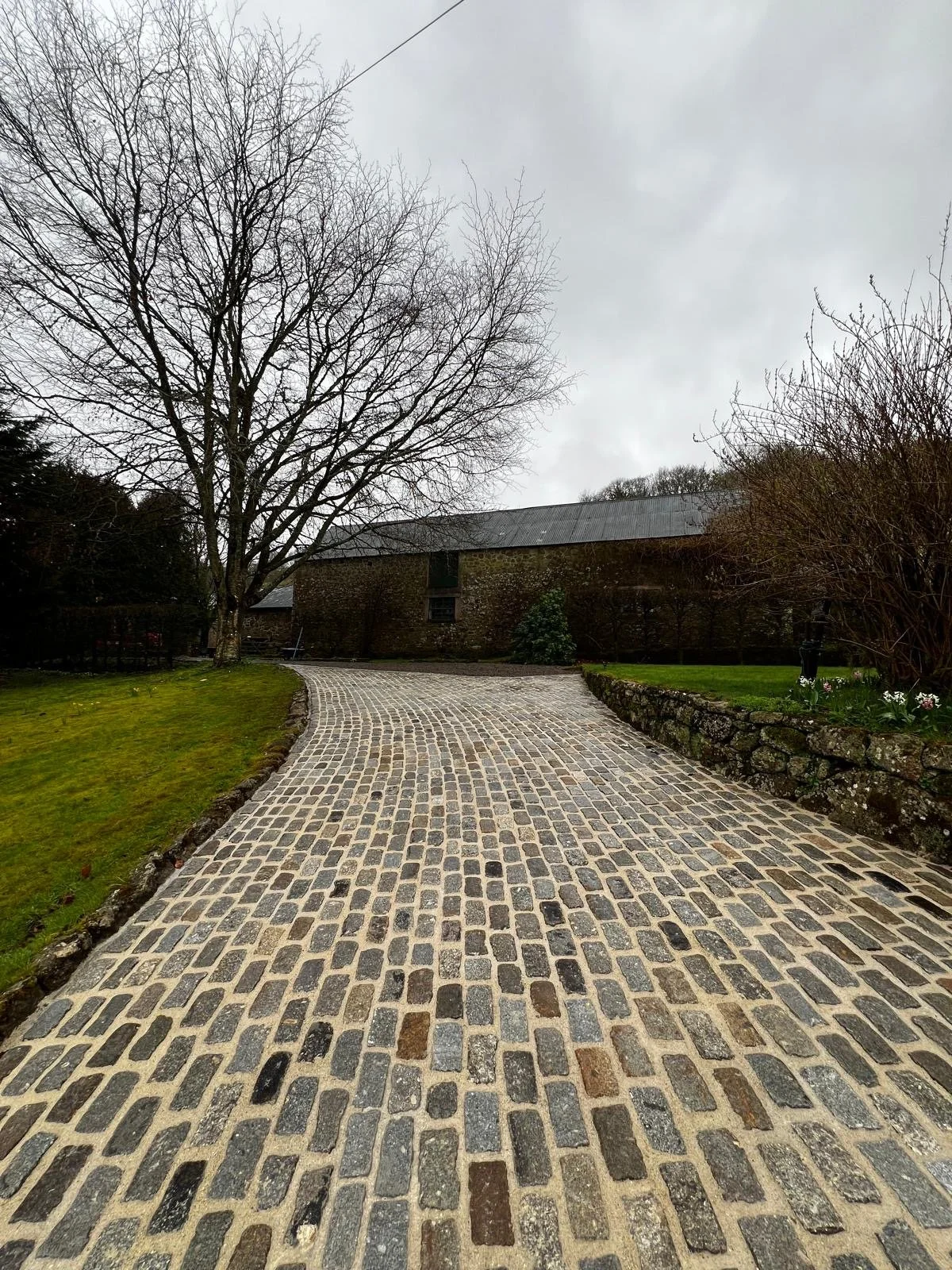 A cobblestone driveway curves toward a rustic brick building with a metal roof, surrounded by a garden with bare trees, bushes, and a small flower bed on a cloudy day.