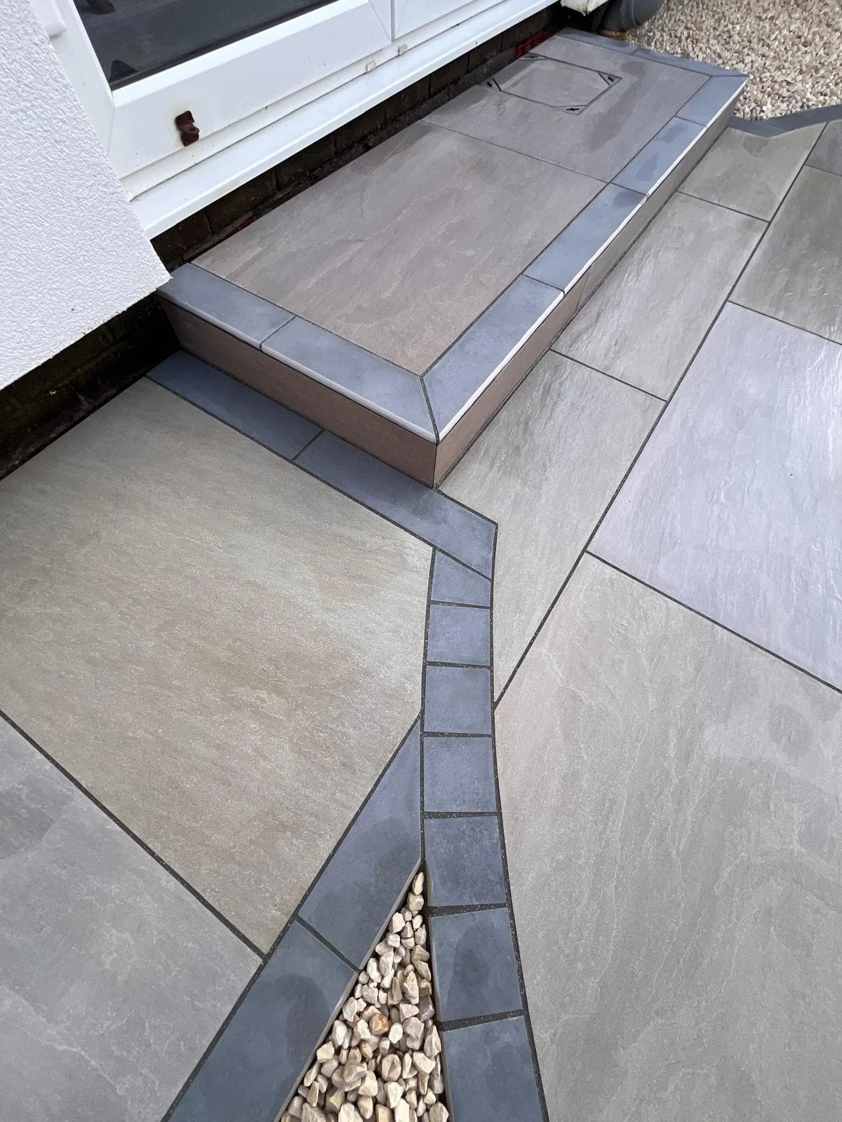 Close-up of a modern outdoor paving area with large rectangular gray tiles and smaller dark gray tiles creating a geometric pattern including a drain cover and a section with small white gravel stones.