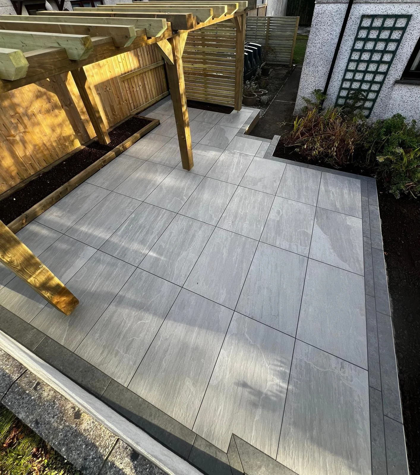 A backyard patio with large gray tiles, wooden fencing, and garden beds.