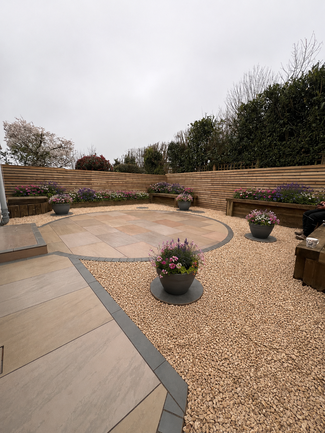 A small, landscaped outdoor patio with flower pots, wooden benches, a wooden privacy fence, and a paved circular area, with trees and shrubs in the background under an overcast sky.