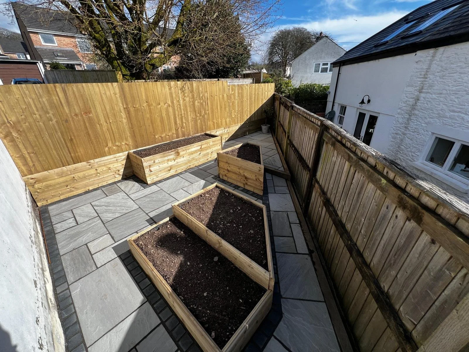 A small outdoor patio with four wooden garden beds filled with soil, surrounded by a tiled floor, enclosed by a wooden fence, with trees and neighboring houses visible in the background.