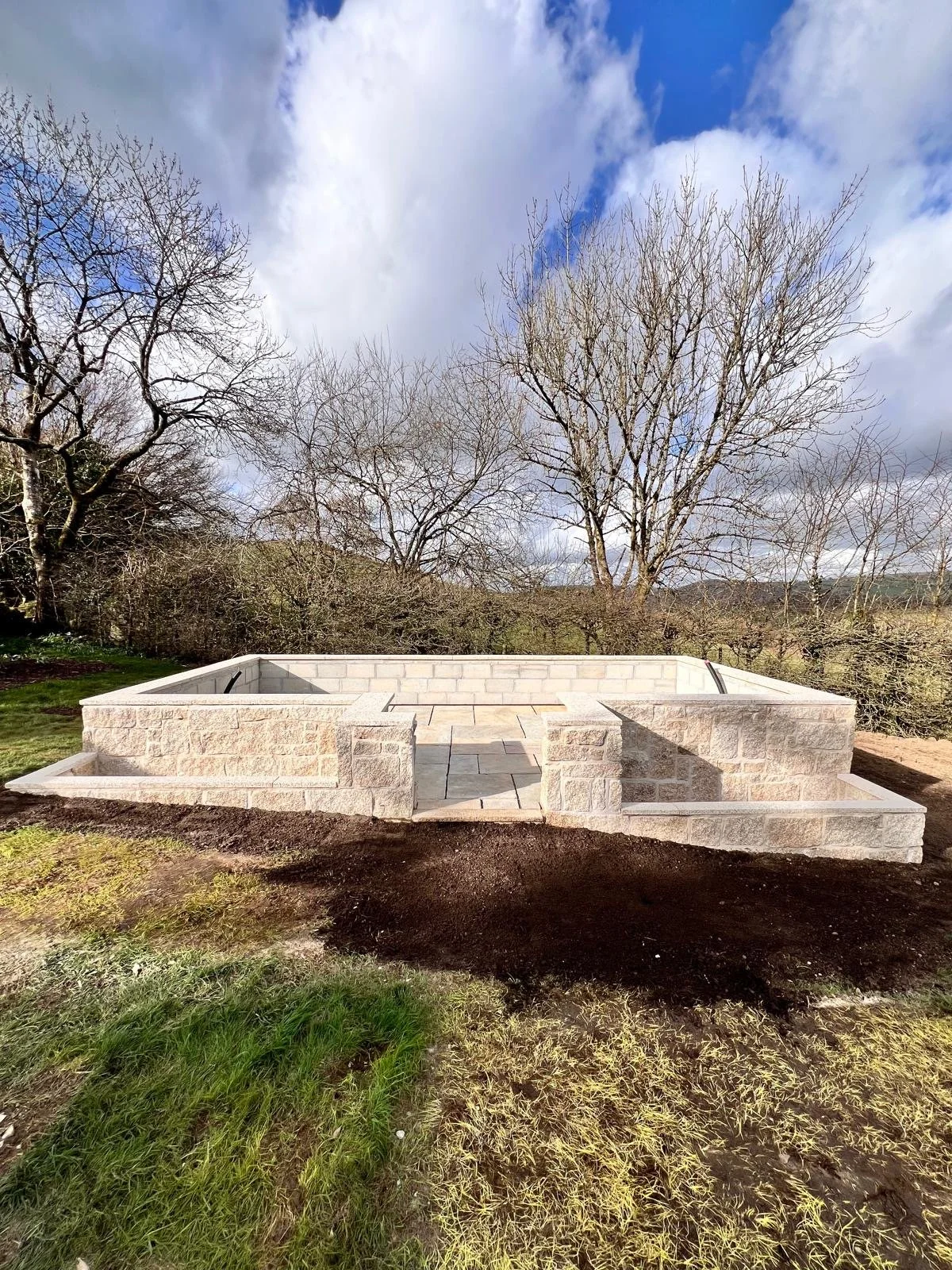 Empty stone hot tub with steps in a backyard, surrounded by grassy and muddy ground, with leafless trees and cloudy sky in the background.