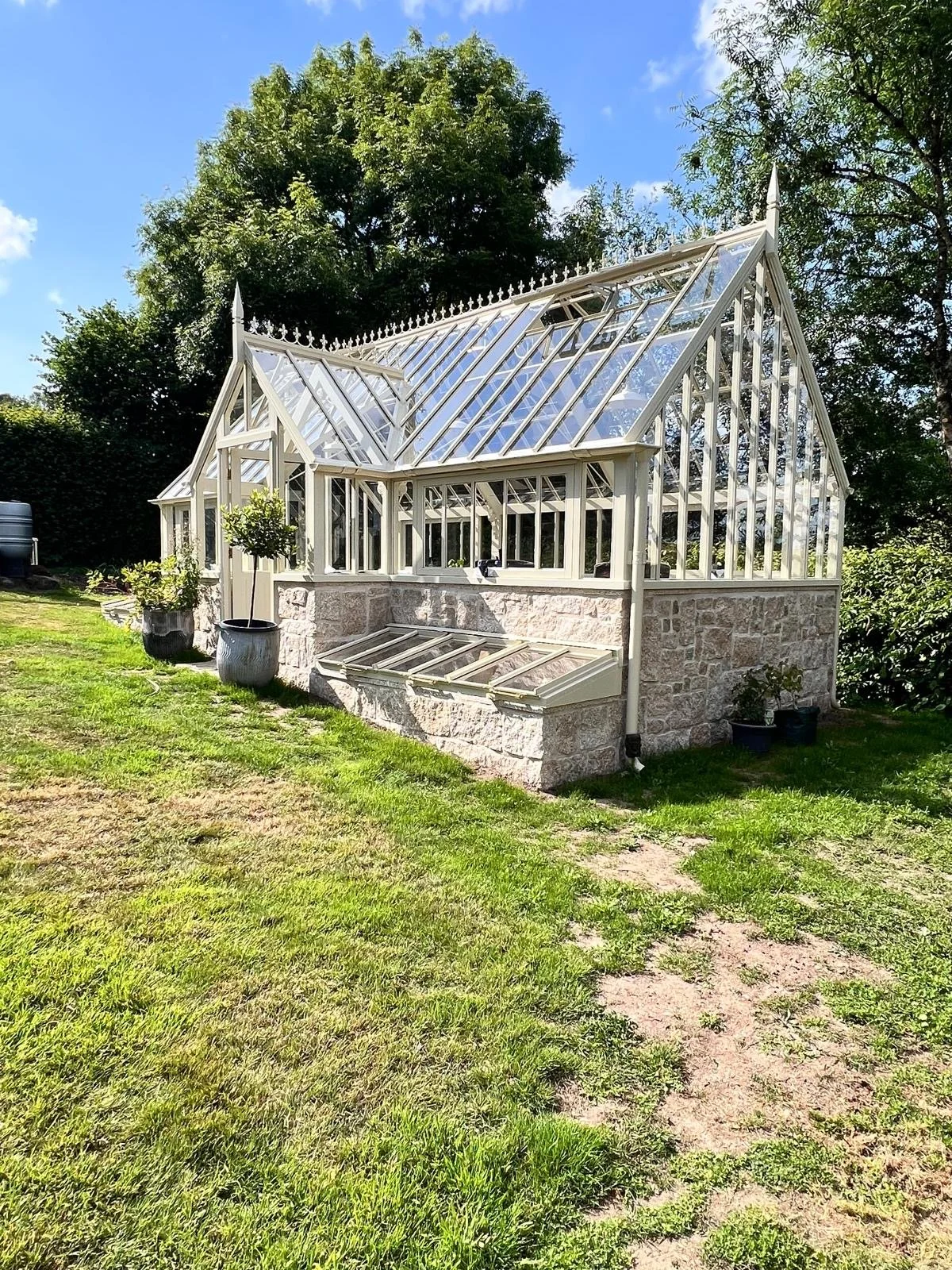 A glass greenhouse with a stone foundation on a grassy lawn, surrounded by trees and blue sky.