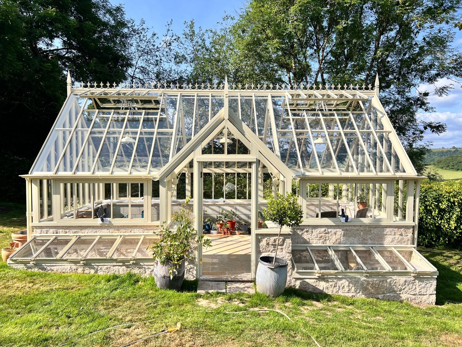 A white greenhouse with glass panels and decorative top finials, surrounded by potted plants and a garden, with trees and sky in the background.