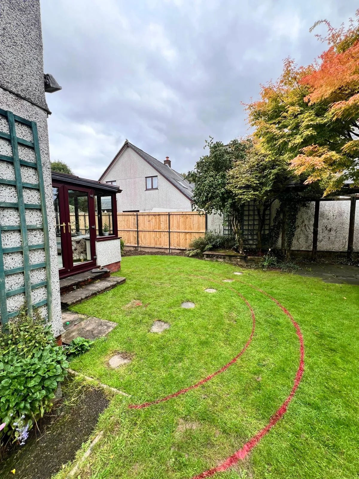 A backyard with green grass, a tree with red and green leaves, a wooden fence, a small shed with a porch and potted plants, and a cloudy sky.