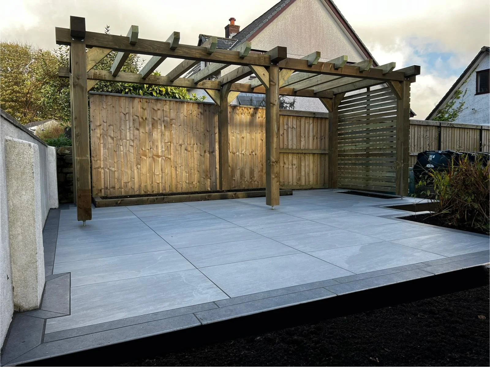 Backyard patio with light gray tiles, wooden pergola frame, and a wooden fence, with plants and neighboring houses in the background.