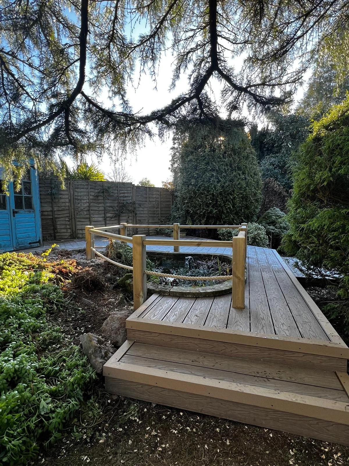 A backyard garden with a wooden deck walkway, a small pond with water fountain, green bushes, and trees, enclosed by a wooden fence and a blue shed.
