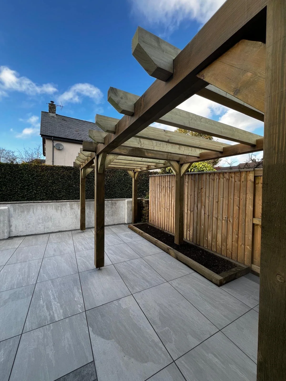 A partially constructed wooden pergola with concrete tiled flooring, a wooden fence, and a garden bed, outdoors under a blue sky with some clouds.