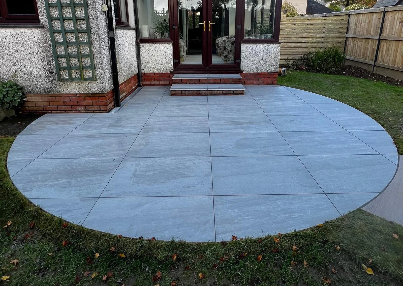 A newly paved circular patio made of large gray tiles, with steps leading to a house entrance, surrounded by green grass and a wooden fence.