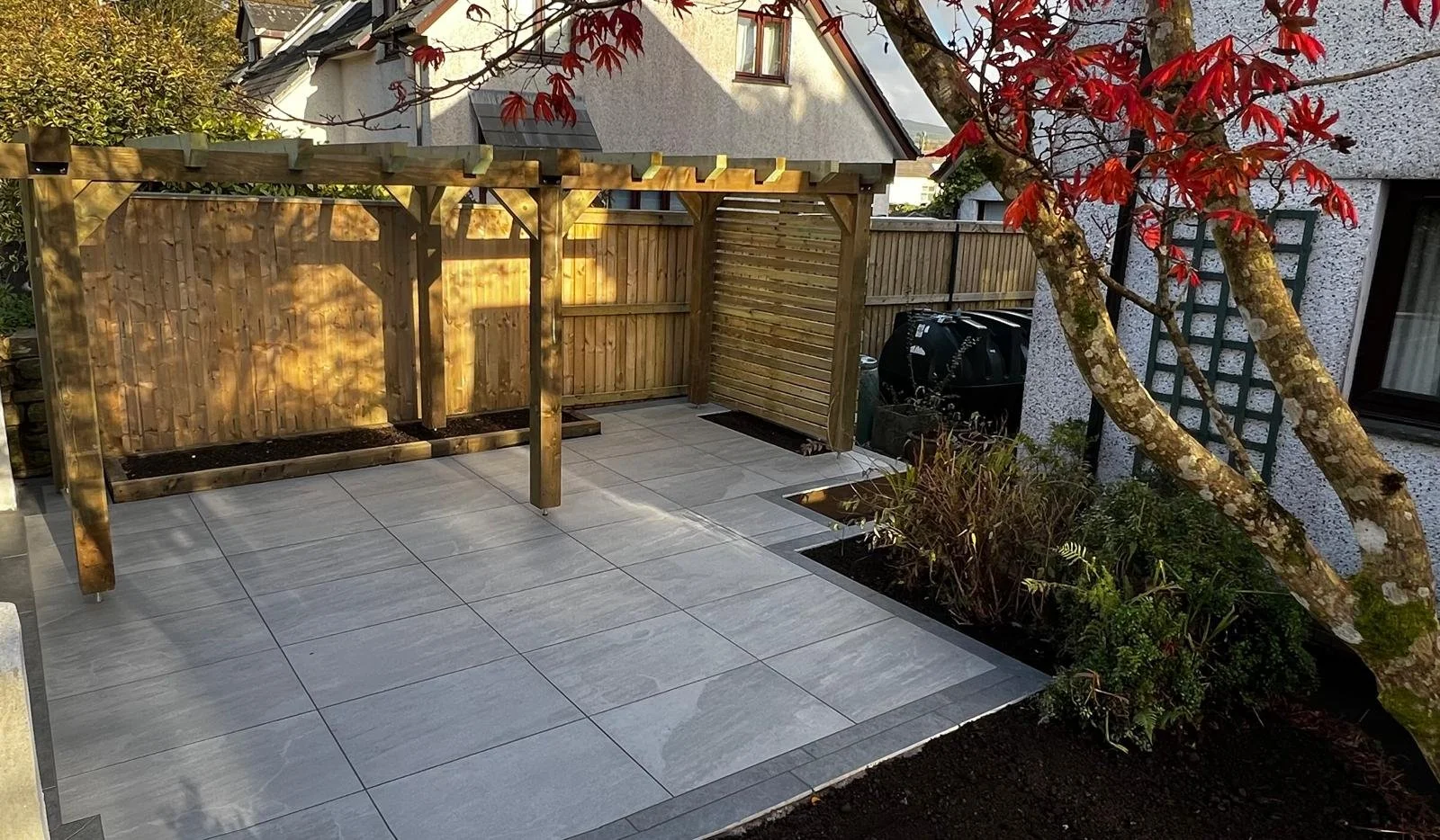 A backyard patio with large gray tiles, a wooden pergola frame, a wooden fence, some plants in garden beds, and a tree with red leaves.