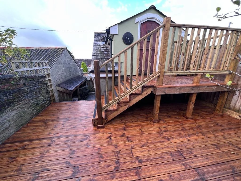Newly built wooden deck with stairs and railing, attached to a small building with a clock, in a backyard with neighboring fences and clear sky.