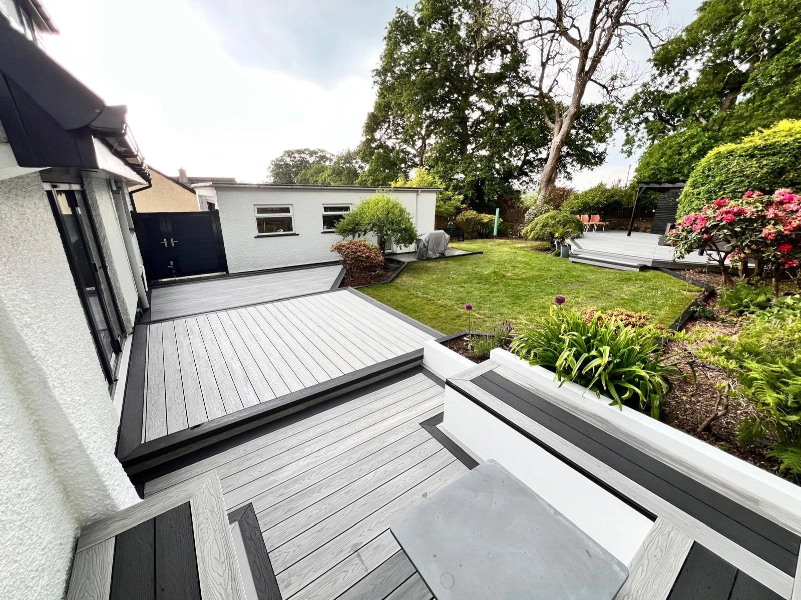View of a backyard patio with gray decking, white walls, green lawn, trees, and flowering plants.