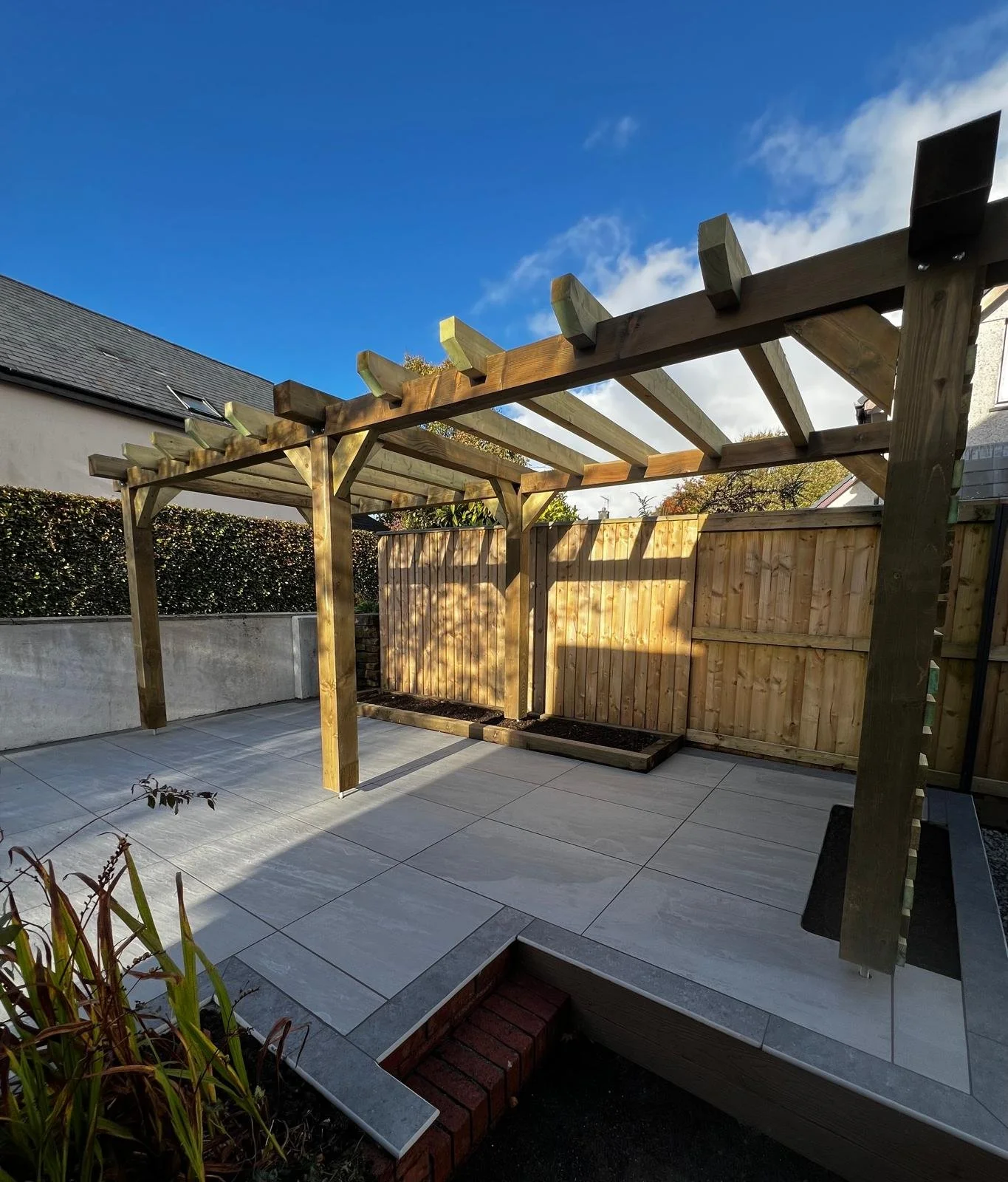 A backyard patio with tiled flooring, wooden pergola framework, and a wooden fence. Sunlight casts shadows, and nearby plants are visible.
