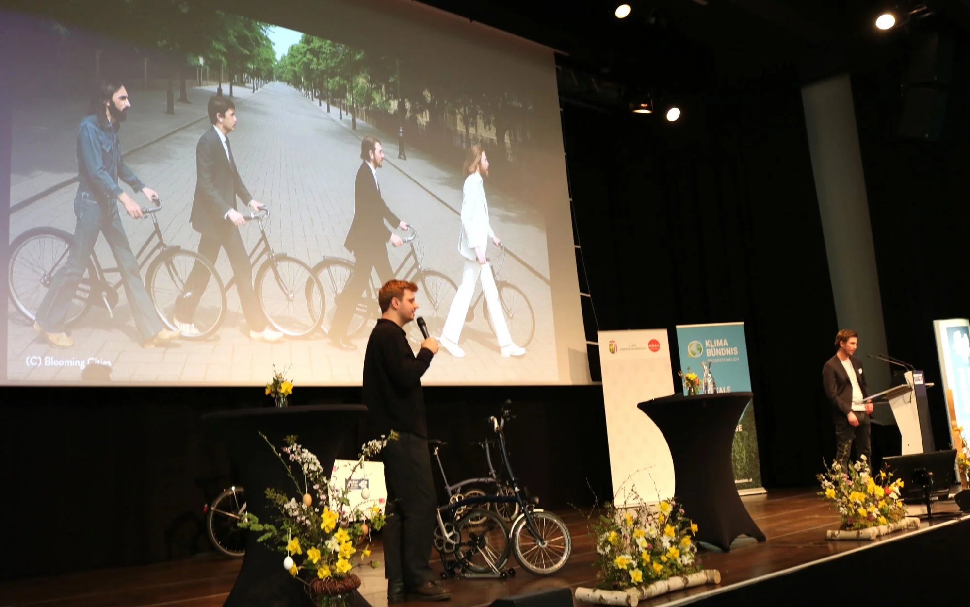 Präsentation auf einer Bühne mit zwei Männern, eines spricht in ein Mikrofon, im Hintergrund eine große Leinwand mit einem Bild von einer Frau und vier Männern, die Fahrrad fahren. Die Bühne ist dekoriert mit Blumen und einer größeren Tafel.