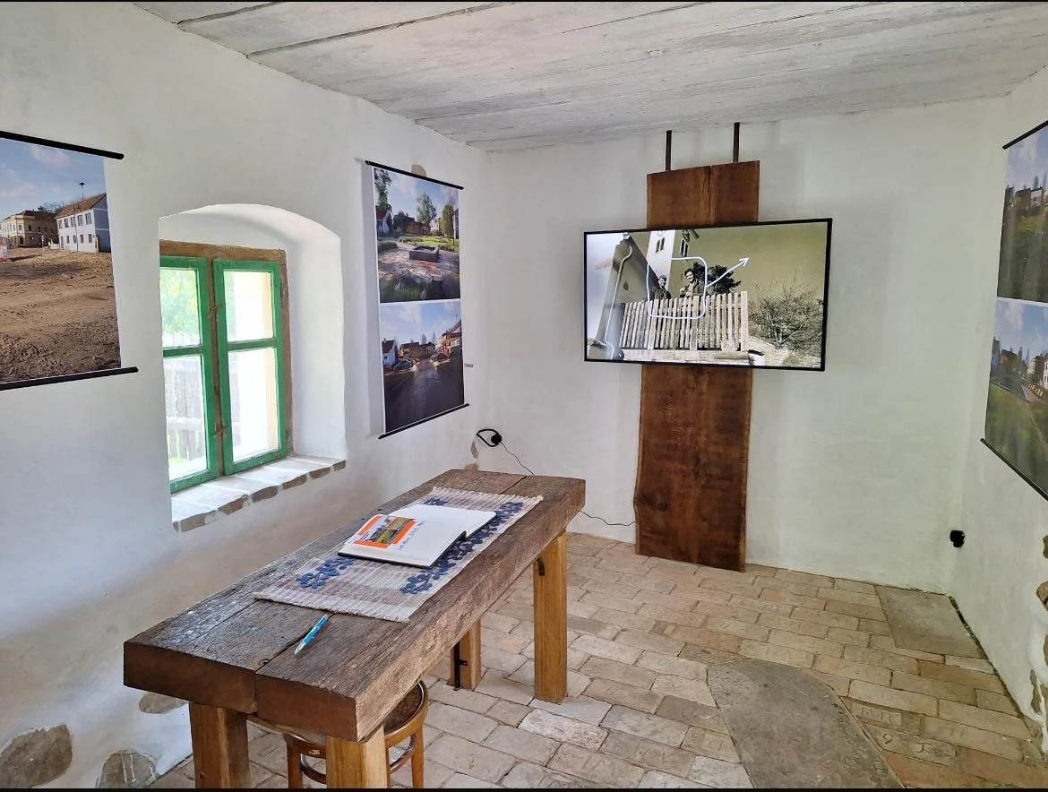 Ein kleines Ausstellungszimmer mit weißen Wänden, einem Holz-Tisch, mehreren an den Wänden hängenden Fotos, einem kleinen Fenster mit grünen Rahmen und einem Fernseher, der eine Skizze eines Gebäudes und Landschaft zeigt.