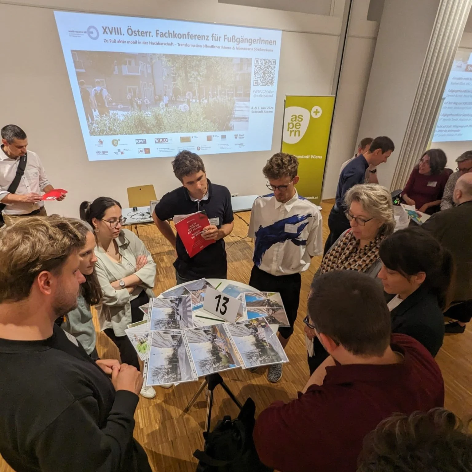 Menschen versammeln sich um einen Tisch mit Fotos und einem Tischnummernzeichen bei einer Konferenz, im Hintergrund ein Bildschirm mit Präsentation und ein gelbes Banner.
