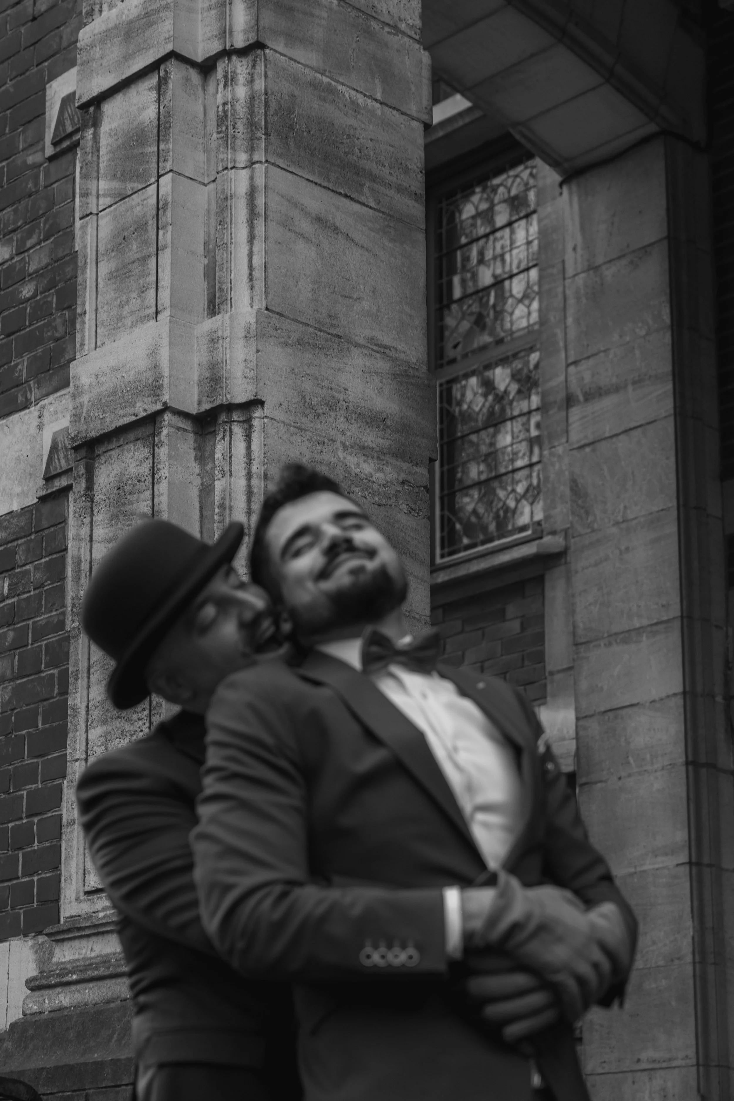 Two men in formal attire, one wearing a bow tie and the other wearing glasses and a bowler hat, share a moment of laughter outside a building with brick and stone architecture.