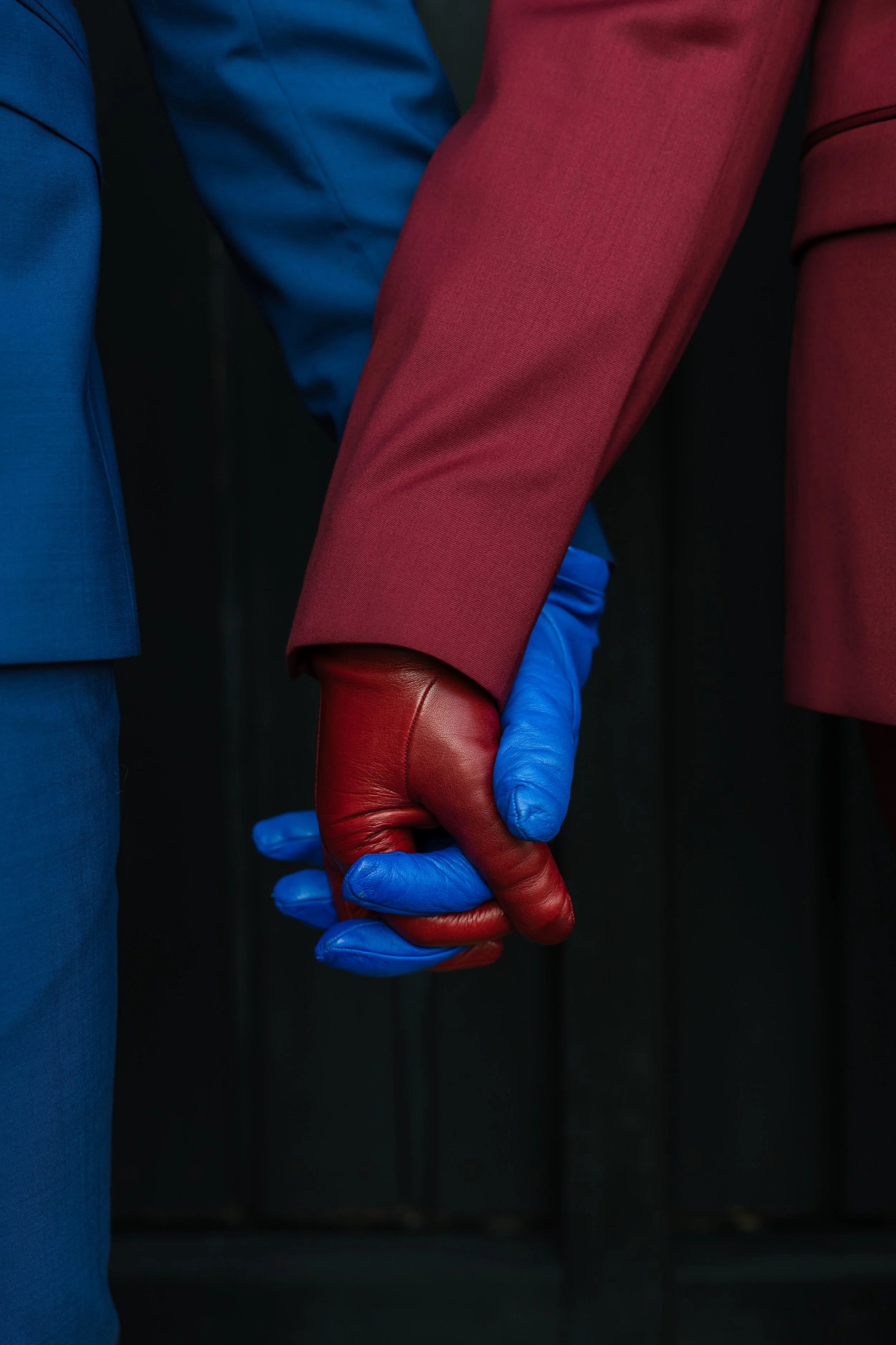 Two people in suits, one in a blue suit and the other in a red suit, holding hands. The person in the red suit is wearing gloves, one red and one blue.