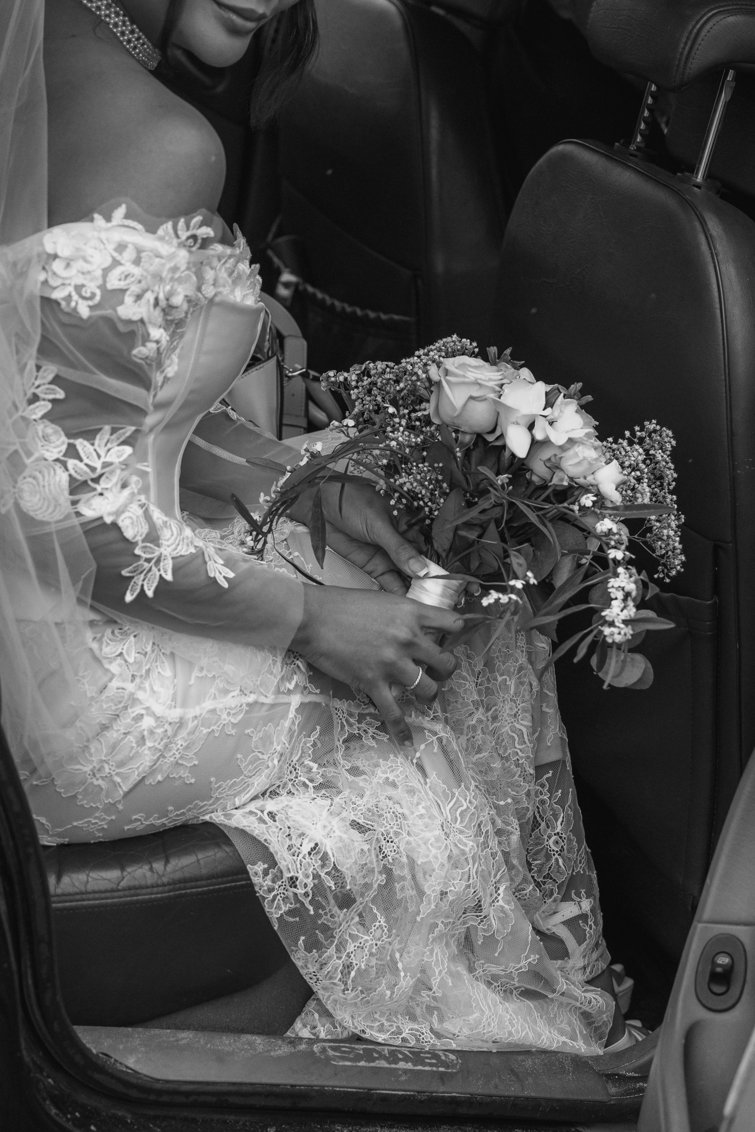 A bride holding a bouquet of flowers sitting in a car, with lace wedding dress and veil partially visible.
