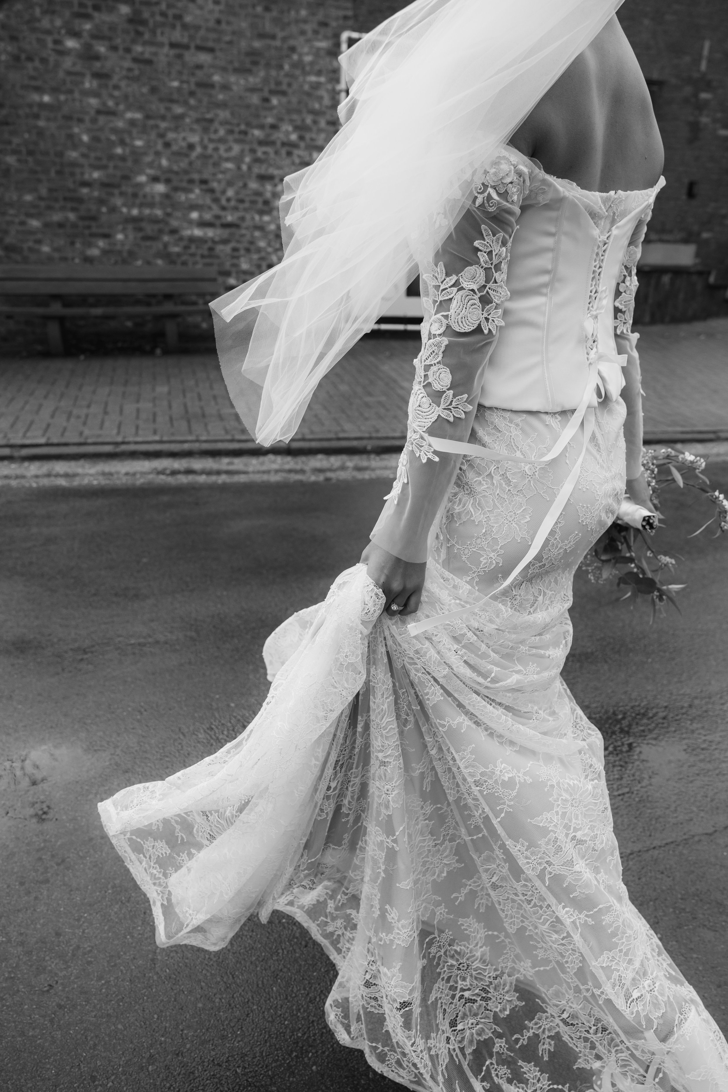 A bride in a lace wedding dress walking outdoors, holding the hem of her dress in one hand, with a veil flowing behind her.